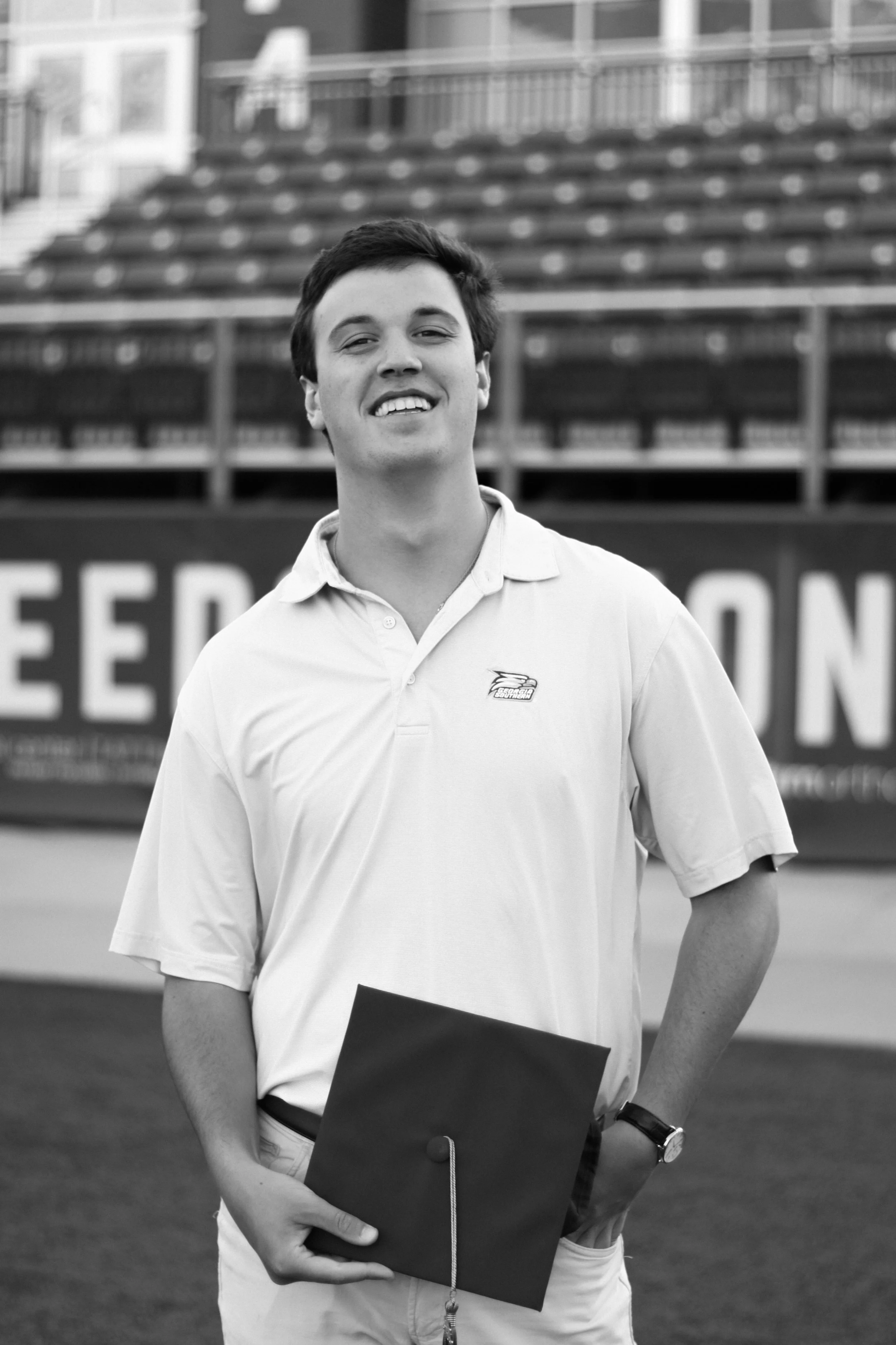 A young man with dark hair and a white polo shirt, holding a dark folder and smiling, standing outdoors in front of stadium bleachers.