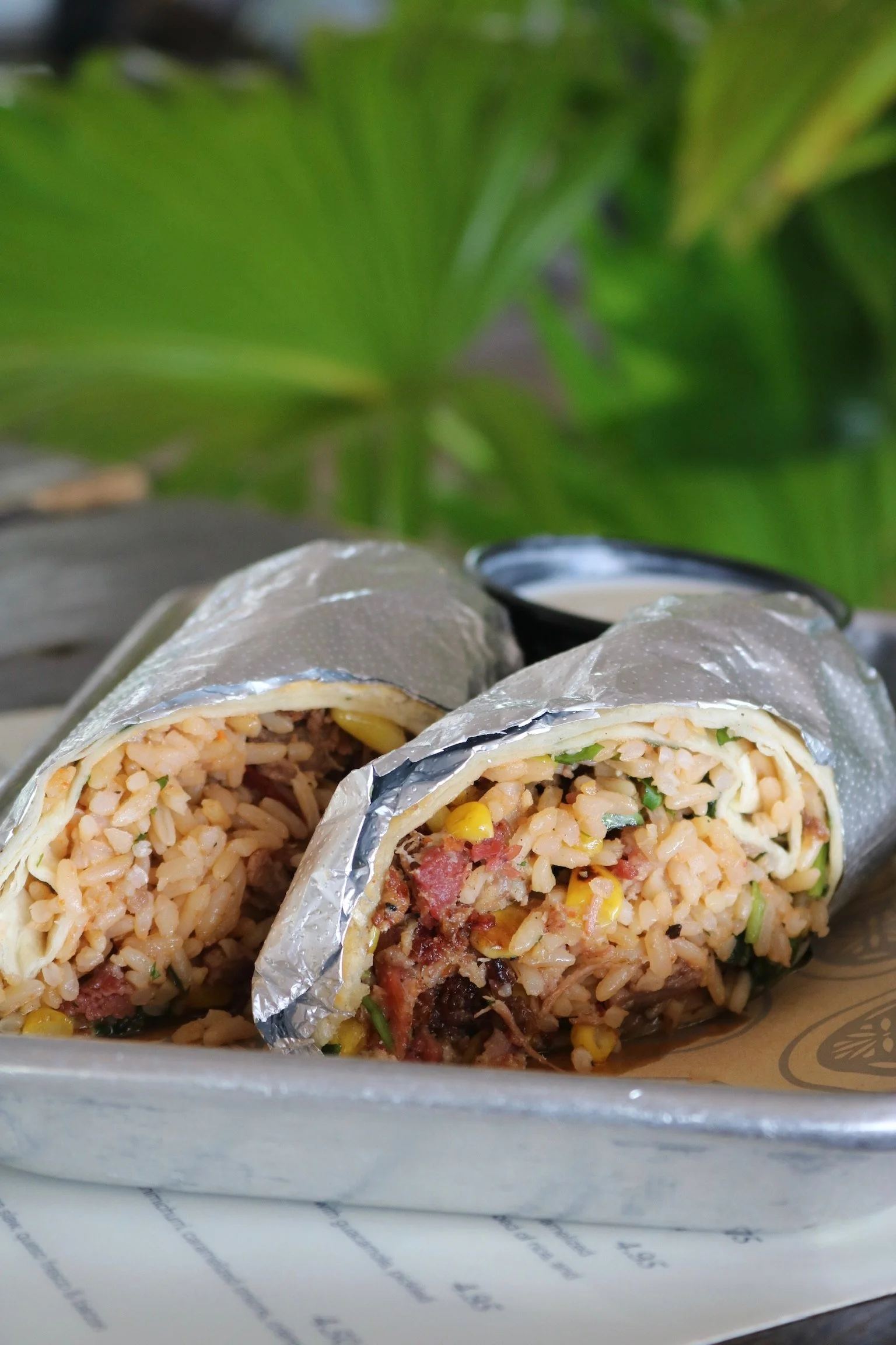 Close-up of two burritos wrapped in foil, filled with rice, beans, corn, and meat, resting on a tray with a green leafy background.