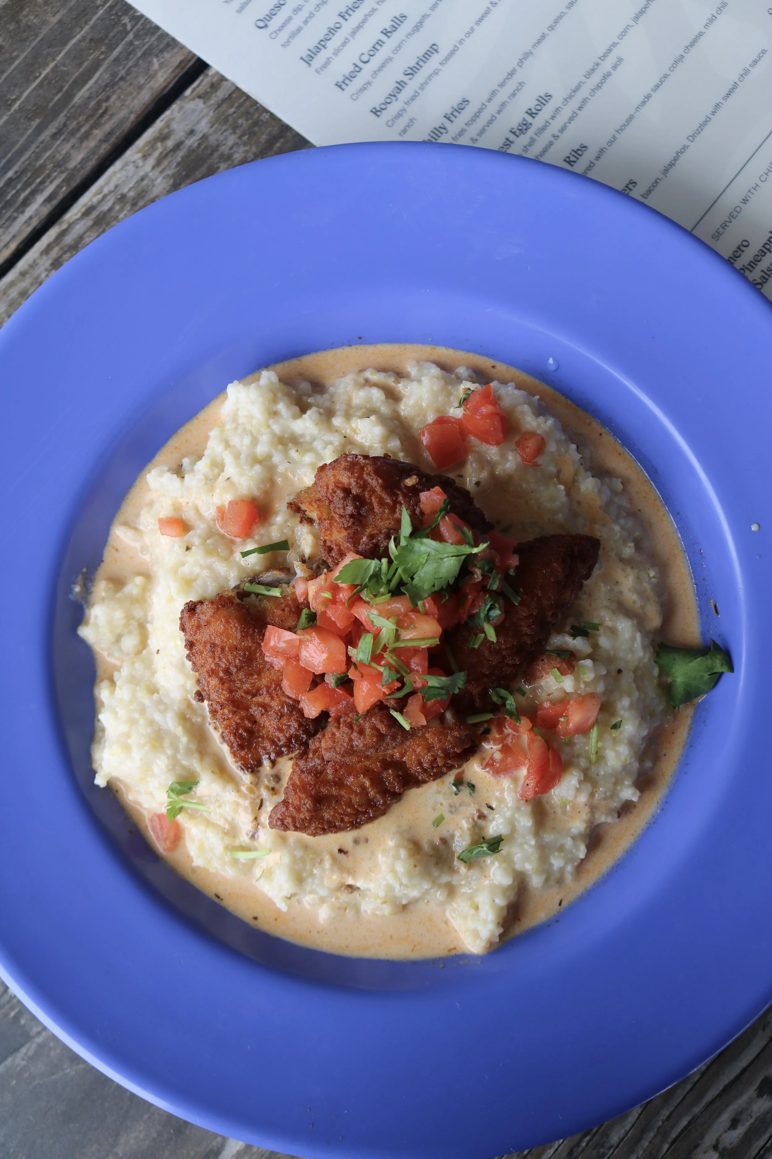 A blue plate of shrimp and grits topped with fried shrimp, diced tomatoes, cilantro, and a creamy sauce on a wooden table