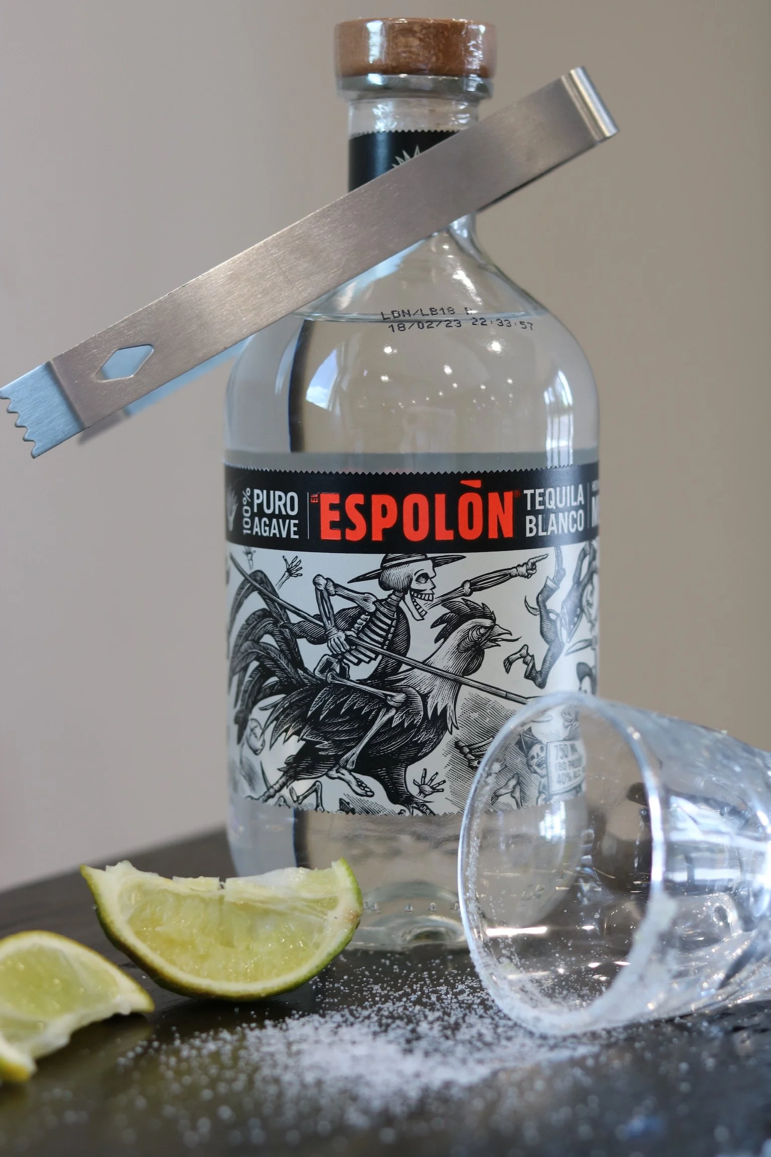 A bottle of Espolon tequila with a skeleton illustration, slices of lime on a dark surface, and an empty glass lying on its side.