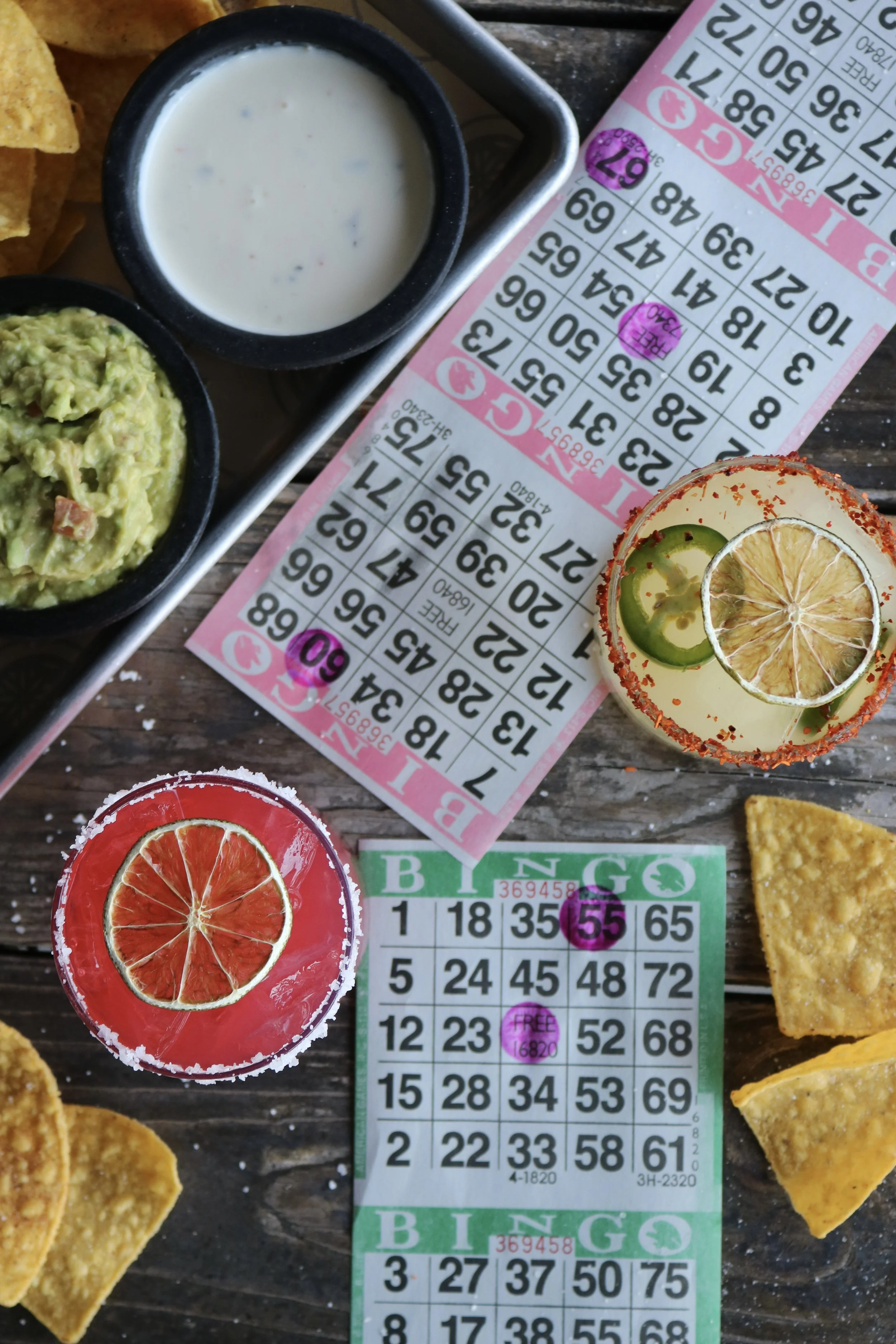 Close-up of a Mexican meal including two shot glasses with salted rim, one containing pink grapefruit juice with a dried grapefruit slice, and the other pink color, alongside a small bowl of guacamole, a small bowl of sour cream or ranch, and tortill