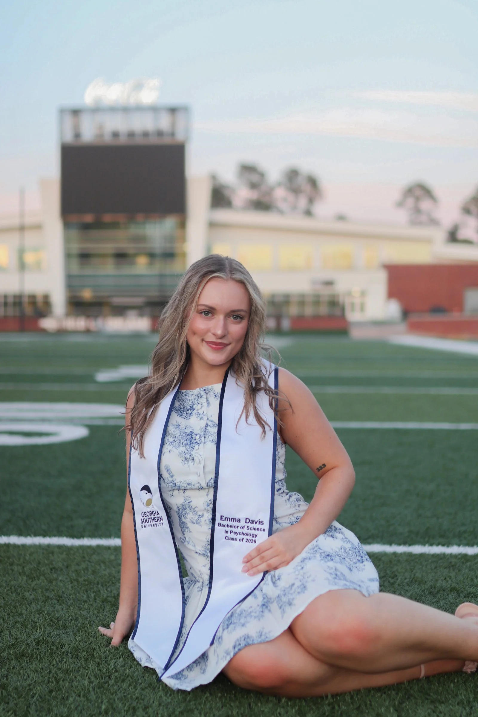 A young woman with long wavy hair sitting on a football field with a university graduation stole around her neck. She is wearing a floral dress, smiling, with a tattoo on her right arm. The background shows a modern building and trees, with a partly 