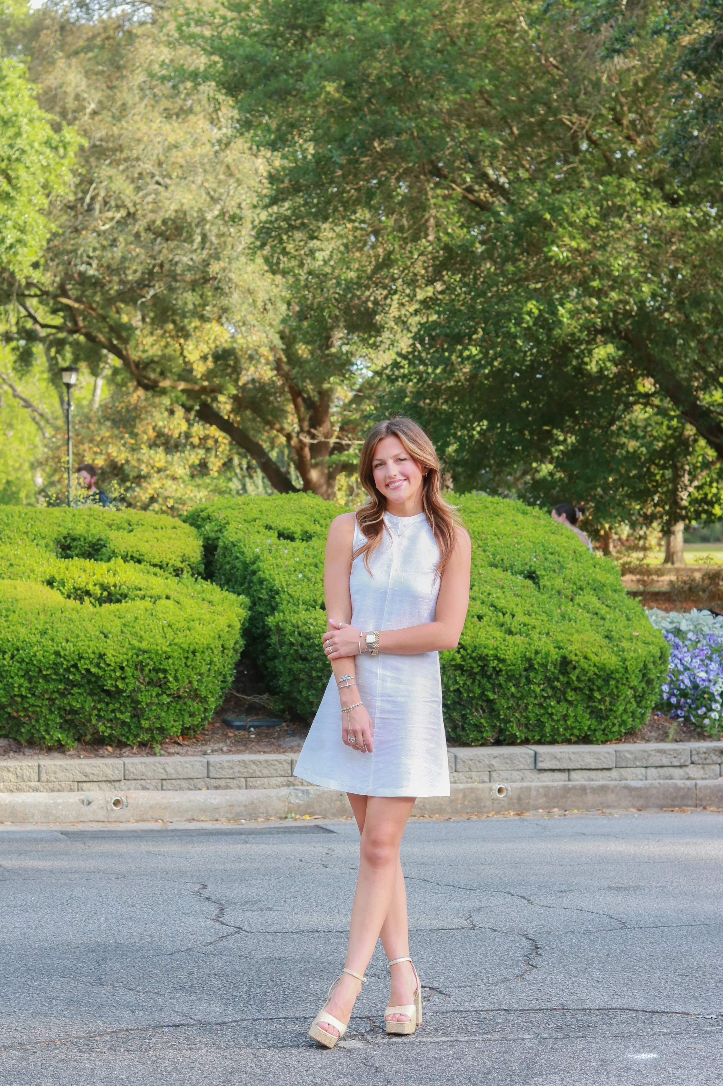 A young woman in a white dress with beige high heels standing outdoors on a paved road with green bushes and tall trees in the background, smiling and crossing her arms.