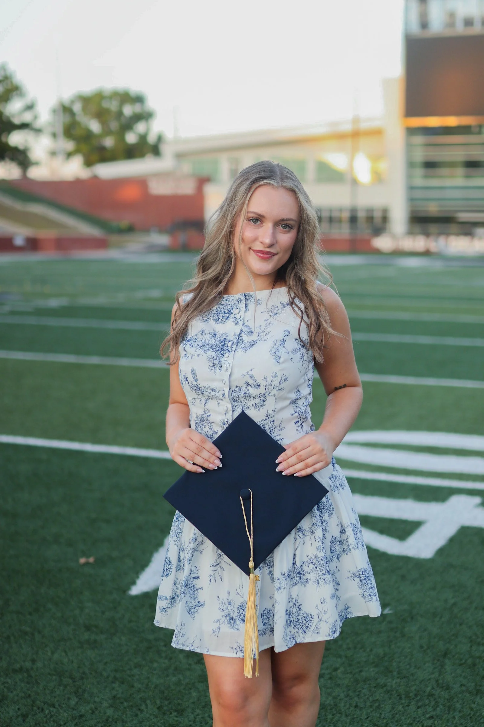 A young woman in a sleeveless white and blue floral dress holding a navy blue graduation cap with a gold tassel, standing on a football field with stadium and trees in the background during sunset.