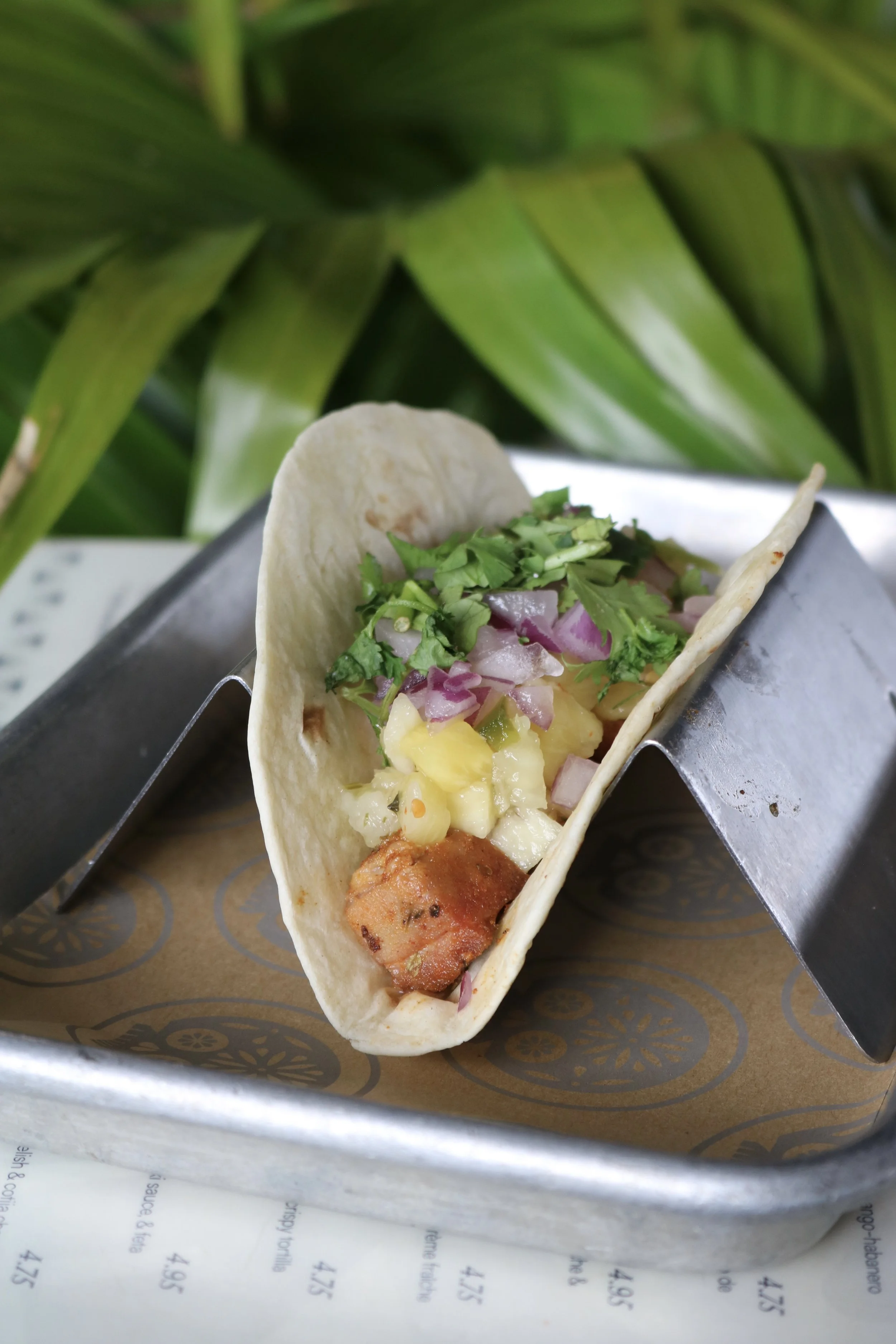 Close-up of a fish taco with toppings, served on a tray with patterned paper and a background of large green leaves.