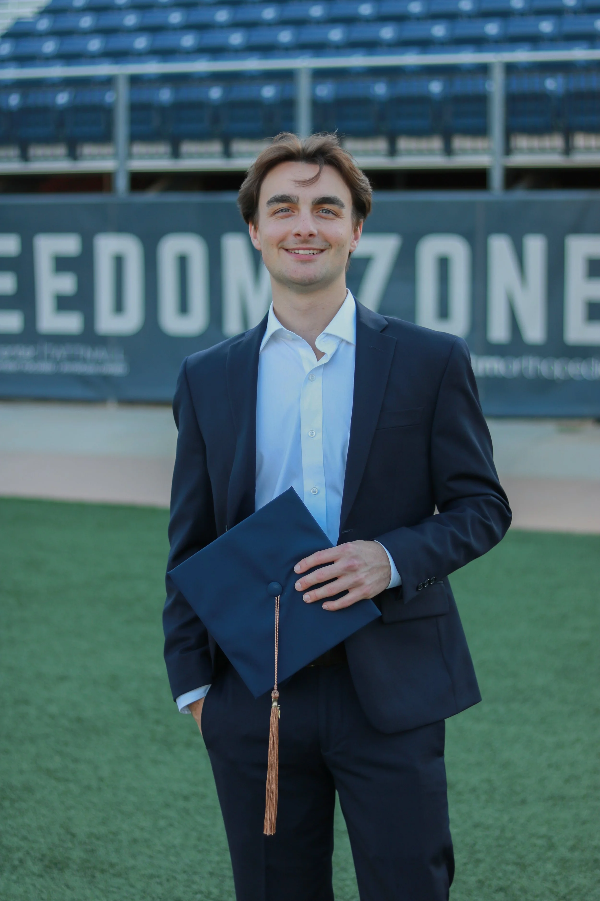 A young man in a navy suit and white shirt holding a navy cap with a brown tassel, standing on a football field with bleachers in the background and a sign that reads 'REDDOMZONE'.