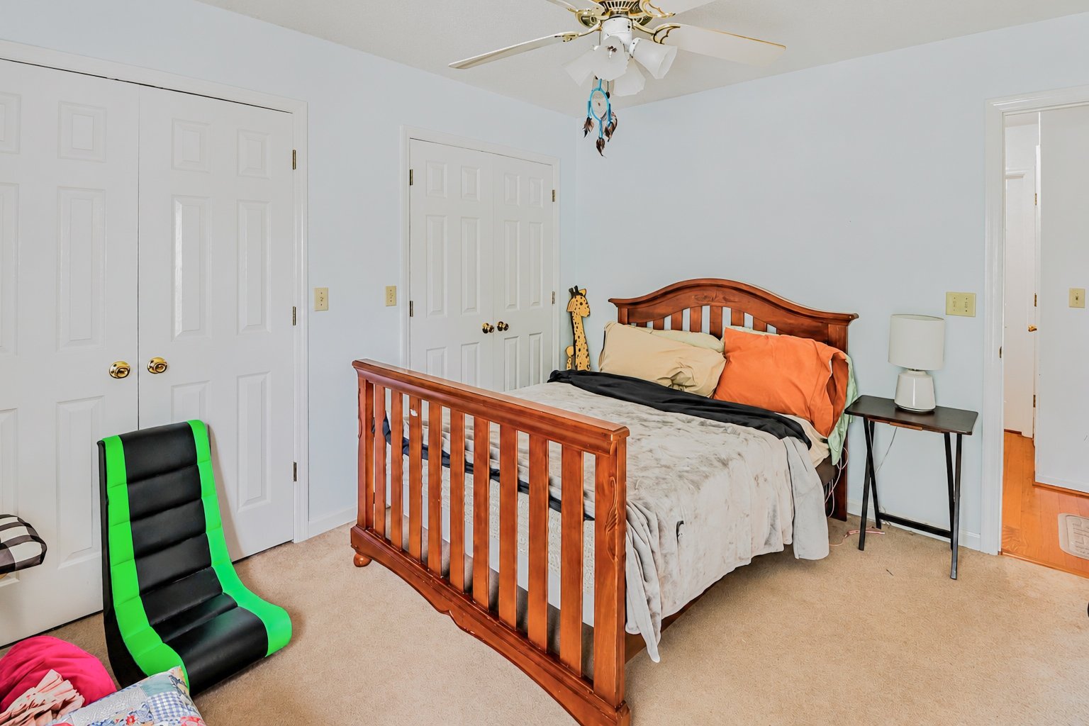 A bedroom with light blue walls, a wooden bed with beige, orange, and black bedding, a small black side table with a white lamp, and a green and black gaming chair in front of a beige carpet.