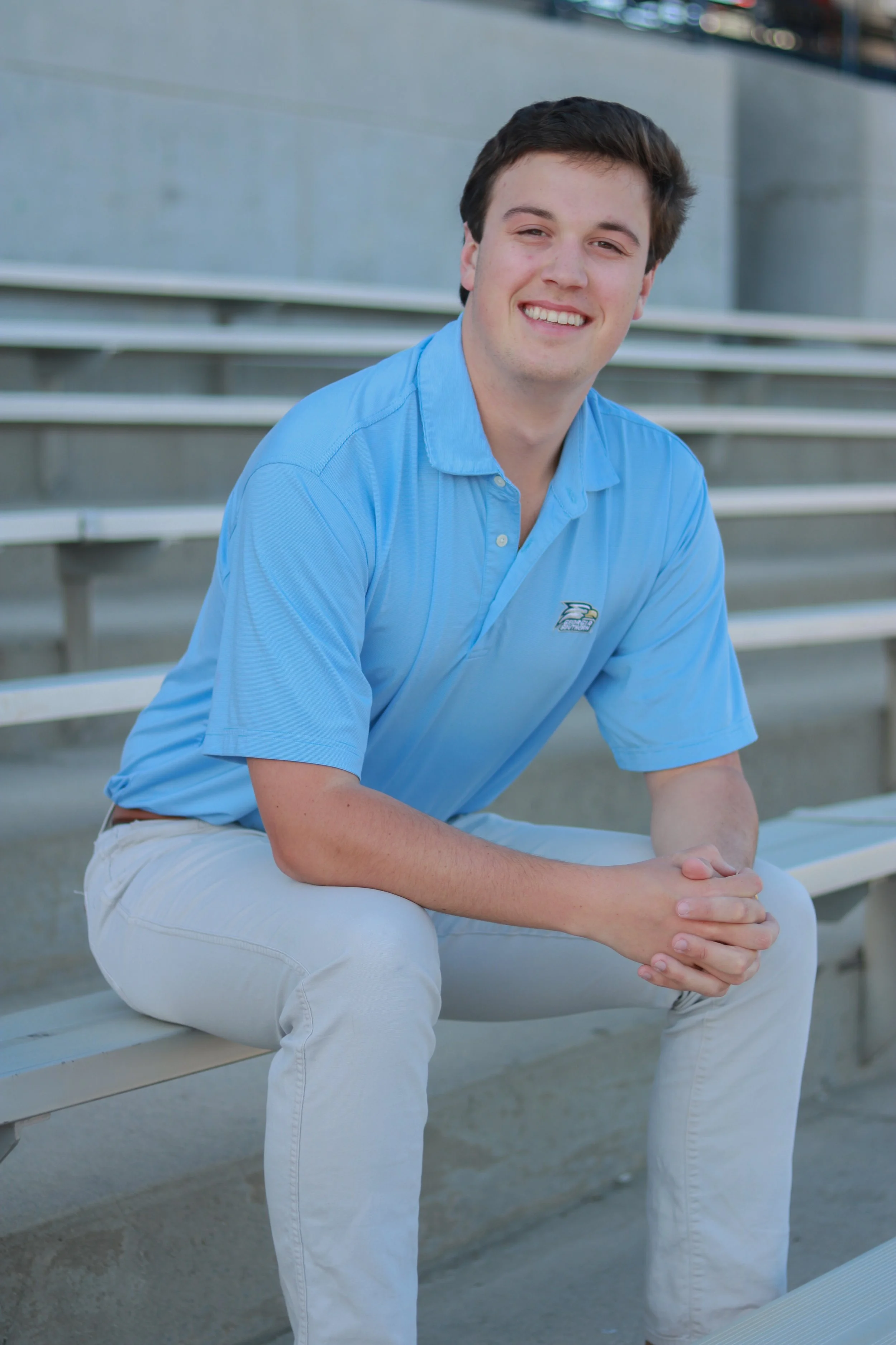 A young man with brown hair and light skin sitting on outdoor bleachers, smiling at the camera. He is wearing a light blue polo shirt and white pants.