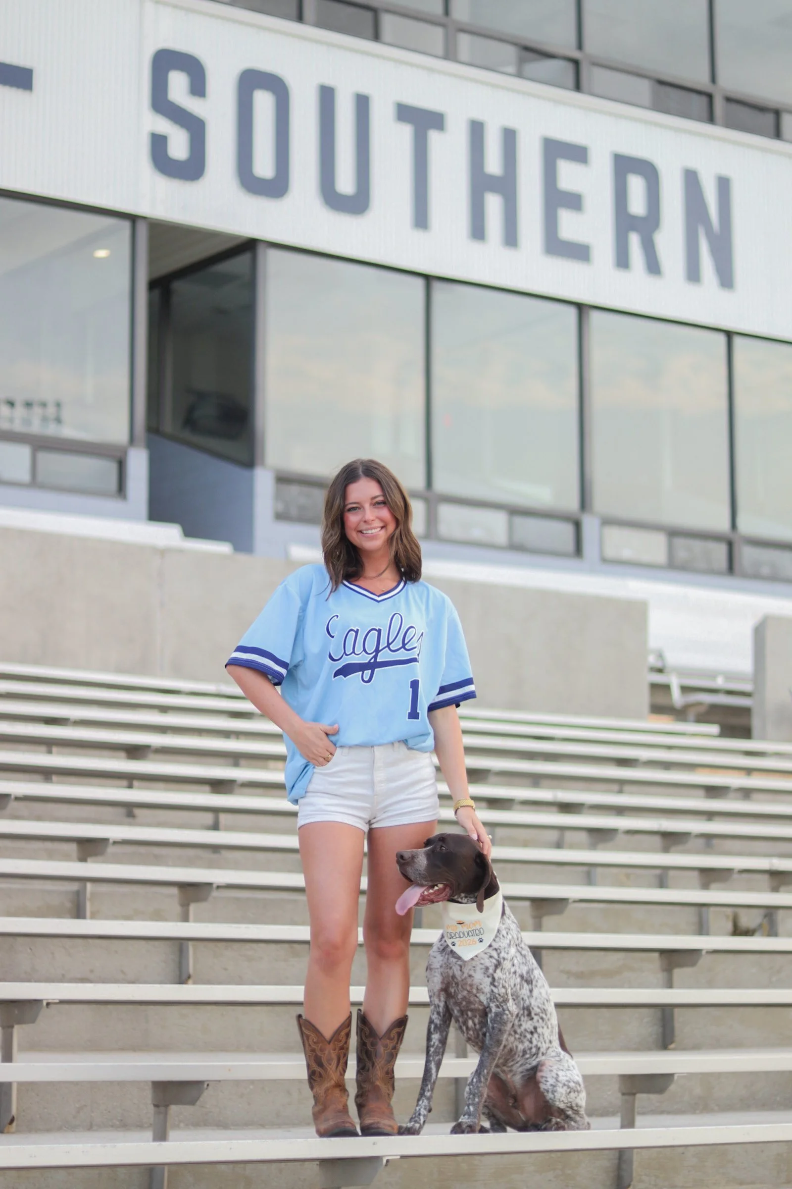 A young woman standing on stadium steps, smiling, wearing a light blue sports jersey and cowboy boots, holding a large dog with a badge around its neck, in front of a modern building with large glass windows and a sign that reads 'SOUTHERN.'