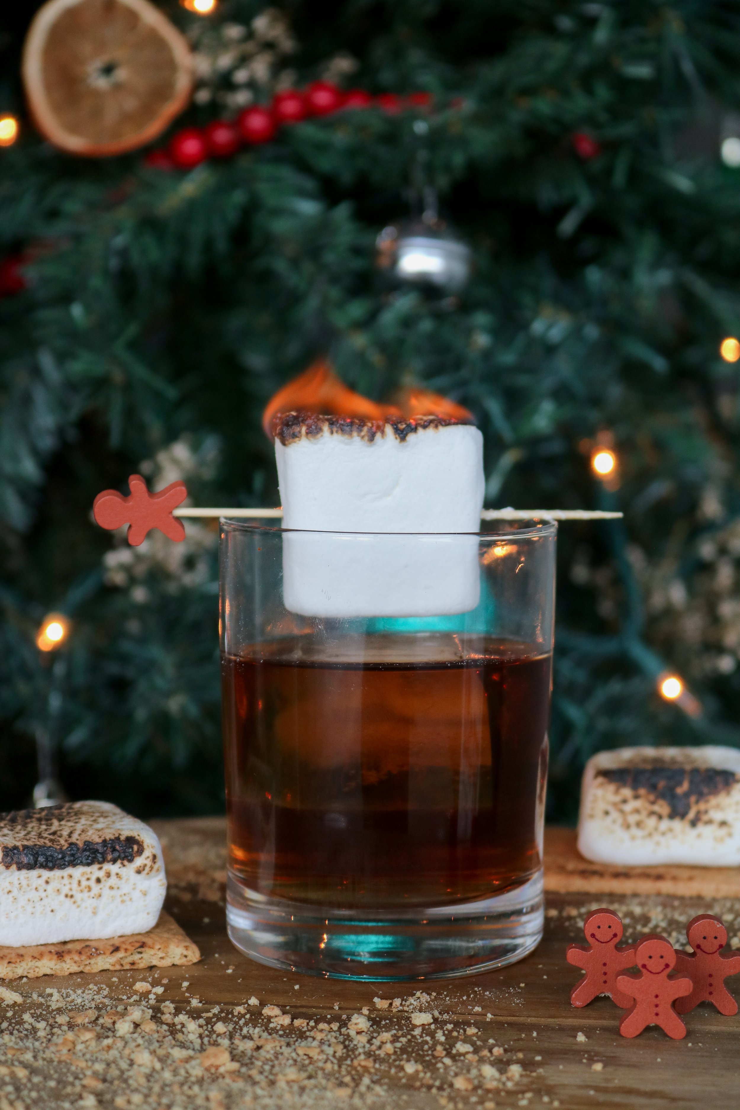 A tall glass of dark beverage with a marshmallow on top, set on a wooden table surrounded by cookies, gingerbread men, and holiday decorations in front of a decorated Christmas tree.