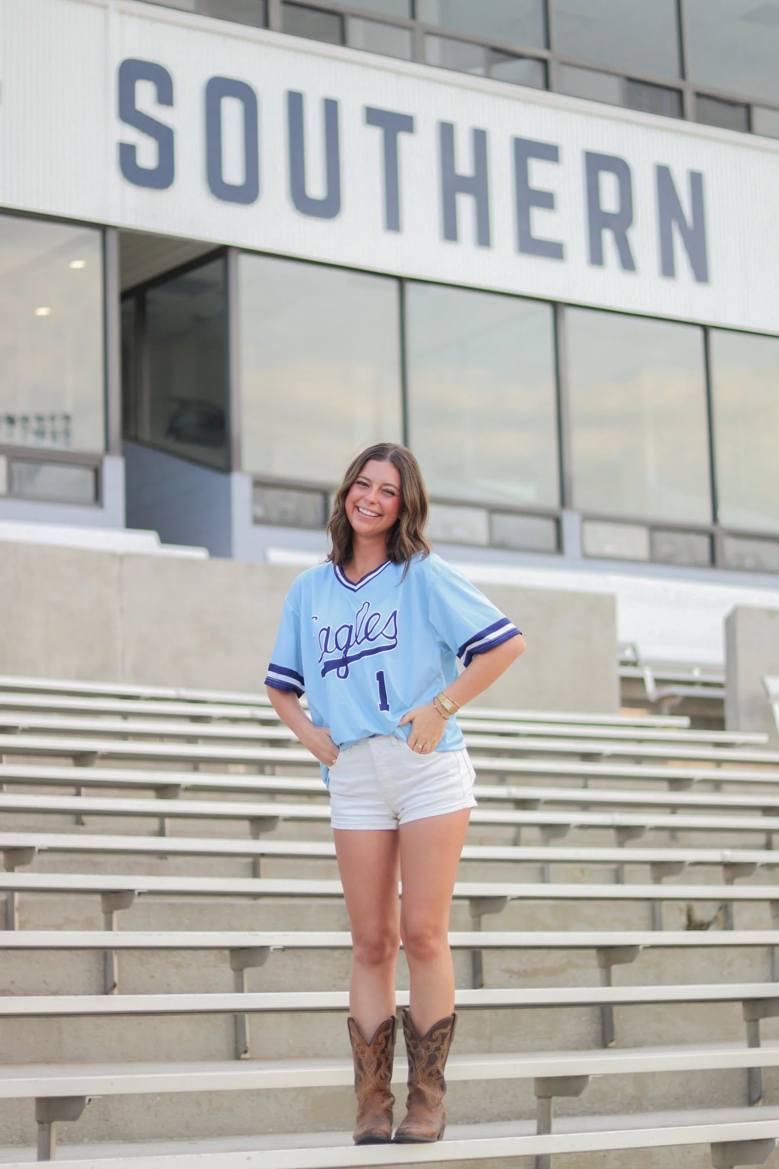 A smiling young woman wearing a light blue baseball jersey with the number 1 on it, white shorts, and cowboy boots, standing on stadium bleacher steps outside a building with the large sign reading 'SOUTHERN' in the background.