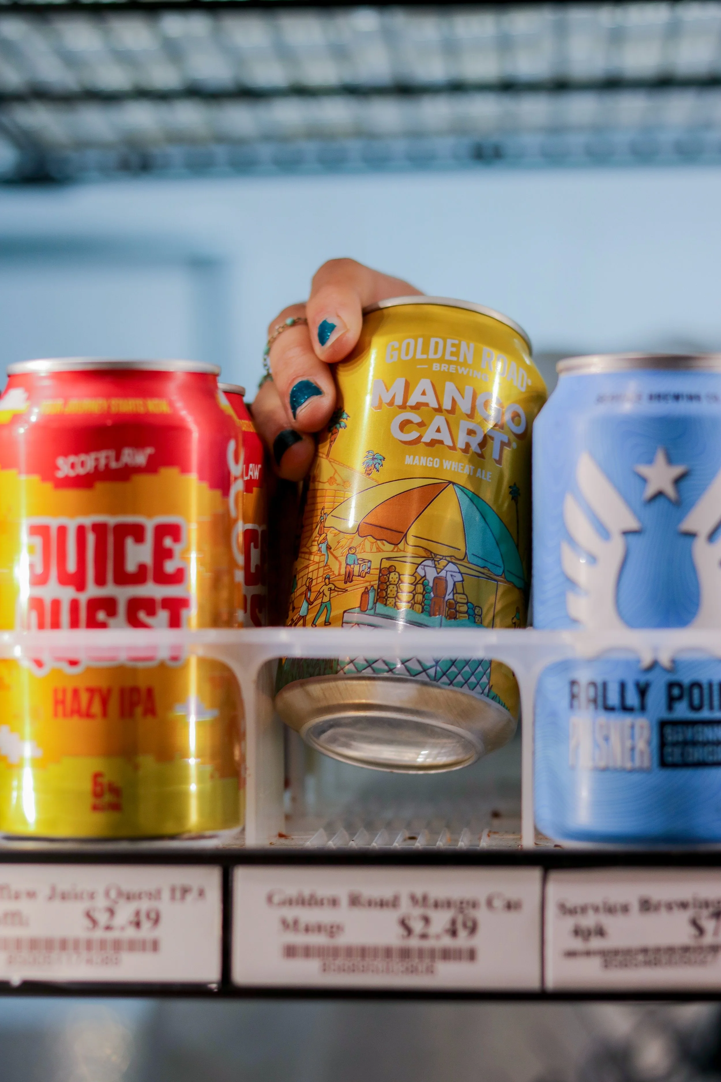 The image shows the inside of a refrigerator door shelf with three cans of beverages, including a yellow can of Mango Cart mango wheat ale, a red can of juice, and a blue can of Rally Point veteran sour. The cans are partially visible and the backgro