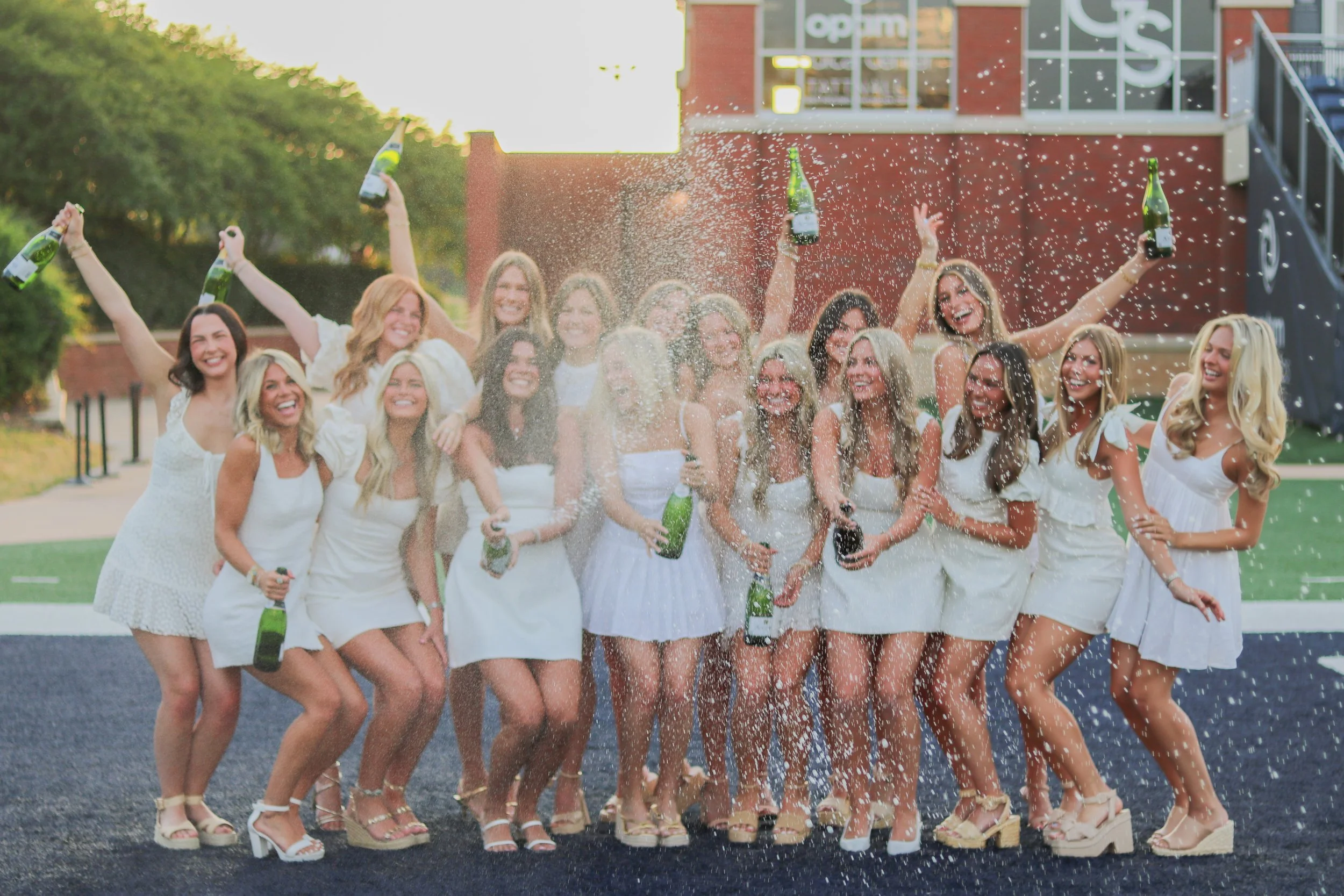 Group of women in white dresses celebrating with champagne bottles and throwing champagne in the air outdoors during sunset.