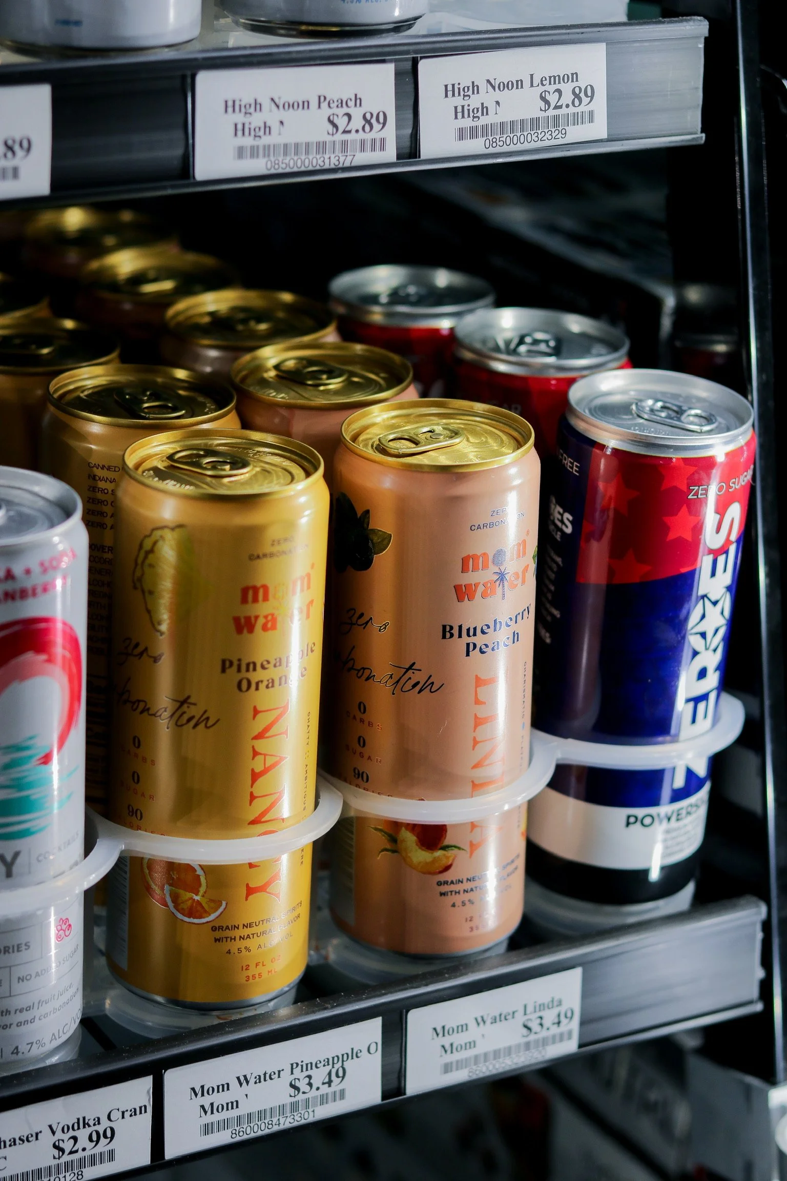 Shelf of canned beverages, including Mom Water flavored drinks in pineapple orange and blueberry peach flavors, and other canned drinks with red and blue packaging.