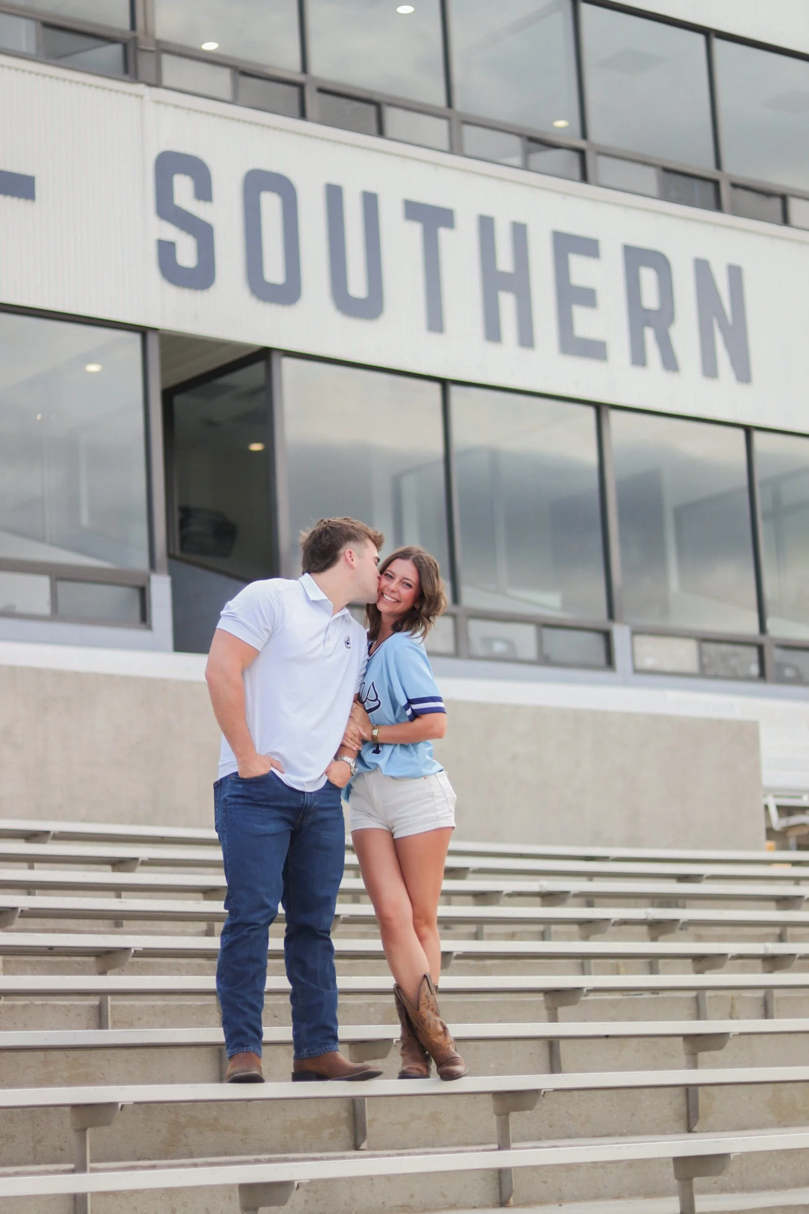 A young man and woman standing on outdoor stadium bleachers in front of a modern building with large glass windows and a sign that reads 'SOUTHERN'. The man is whispering into the woman's ear, and she is smiling.