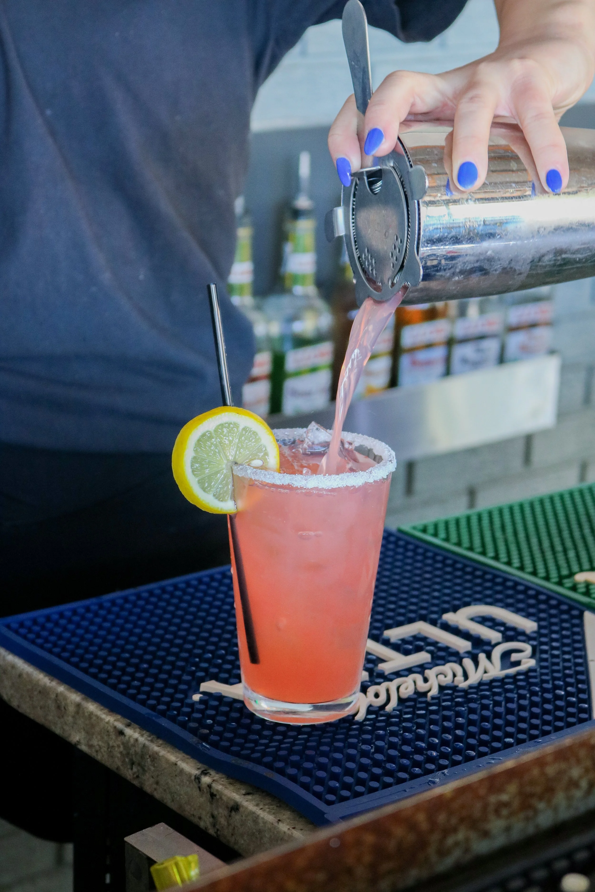 A bartender pouring pink cocktail into a glass with a salted rim, a lemon wedge garnish, and a black straw on a bar counter with a blue mat.