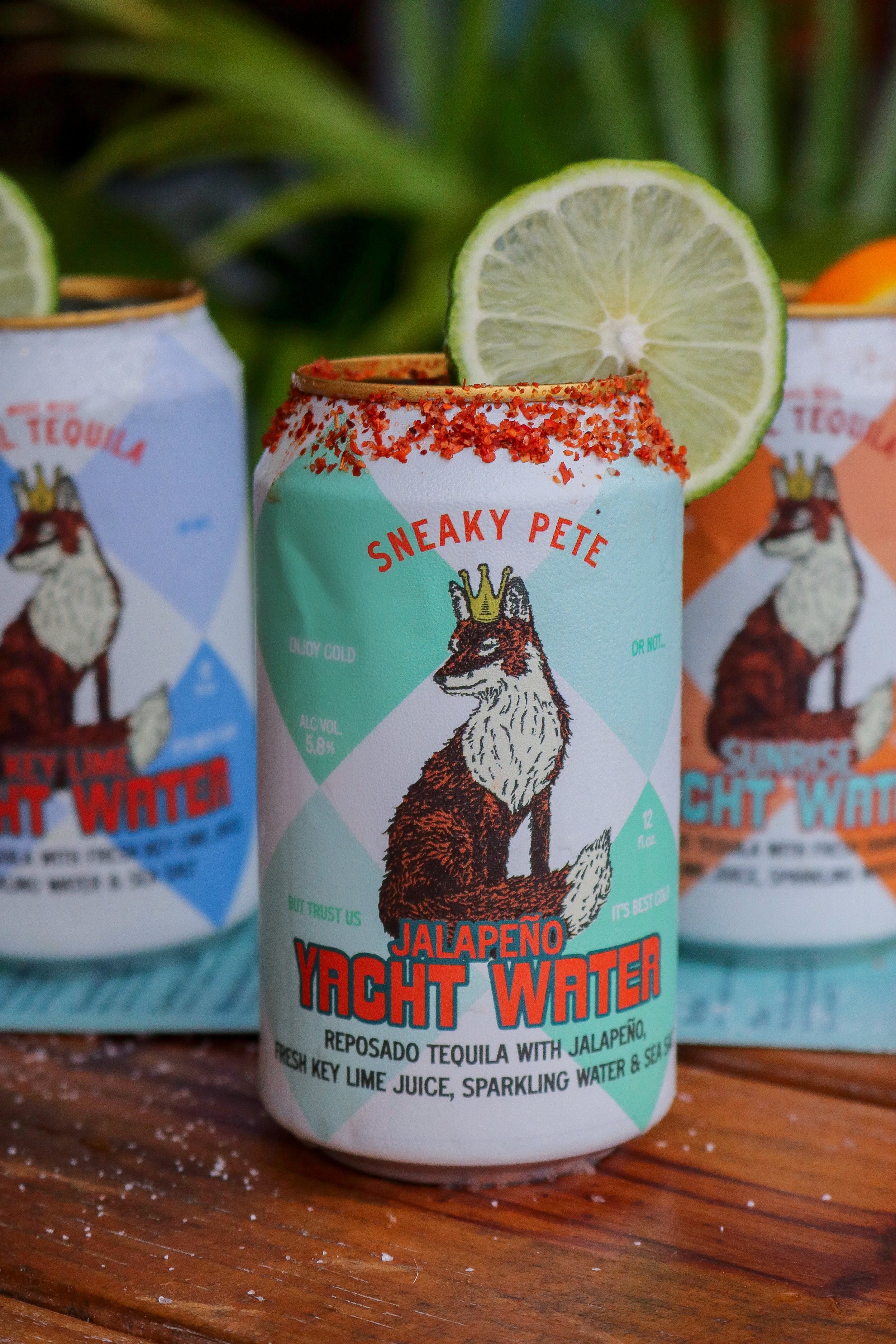 A can of Jalapeño Yacht Water with a slice of lime on top, featuring a cartoon fox wearing a crown on the label, placed on a wooden surface with a blurred background of leaves.