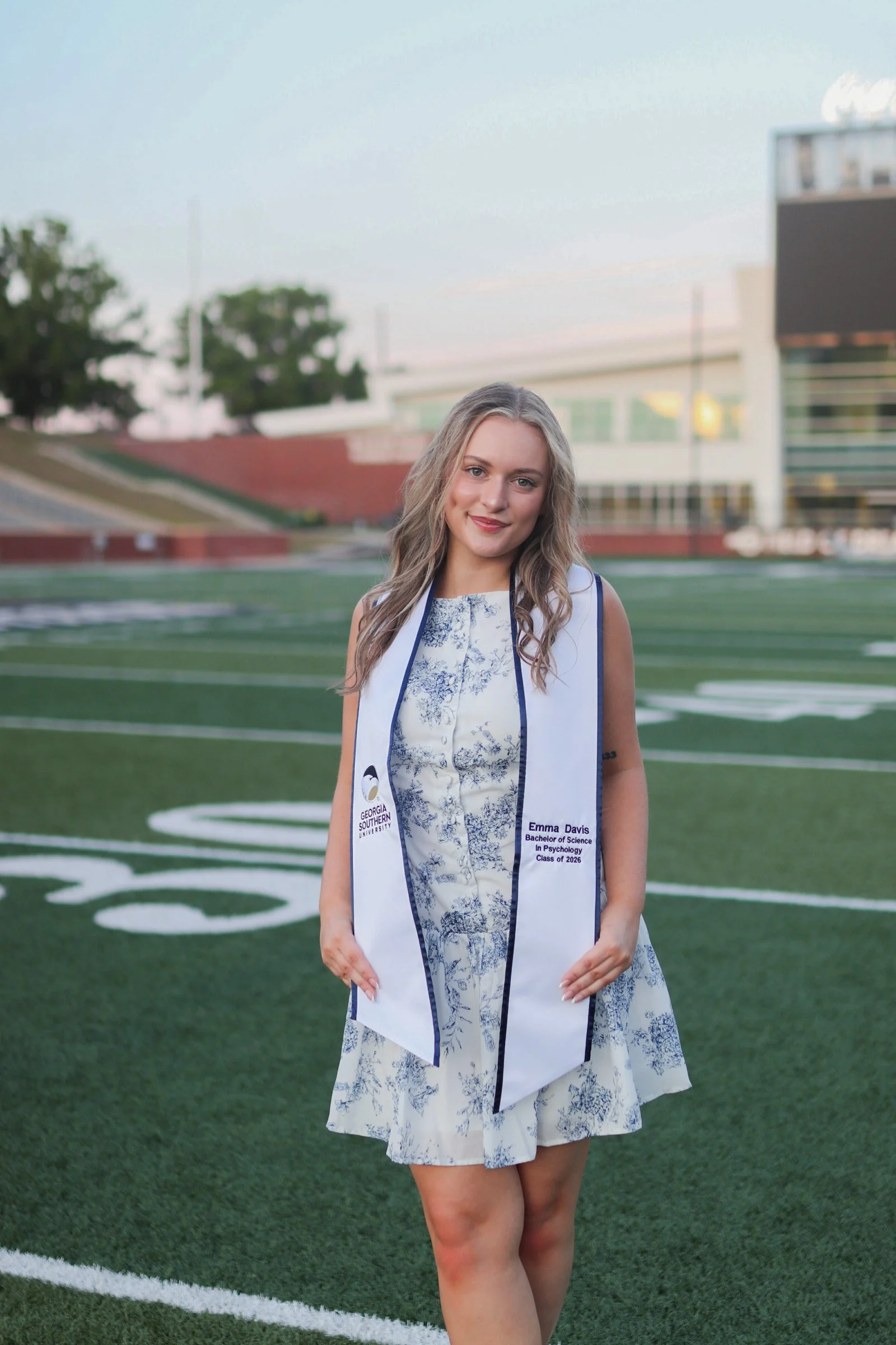A young woman in a floral dress stands on a sports field, wearing a white and blue sash that has her name and academic details. She has blond, wavy hair and a slight smile. In the background, there are empty stadium seats and trees, indicating an out