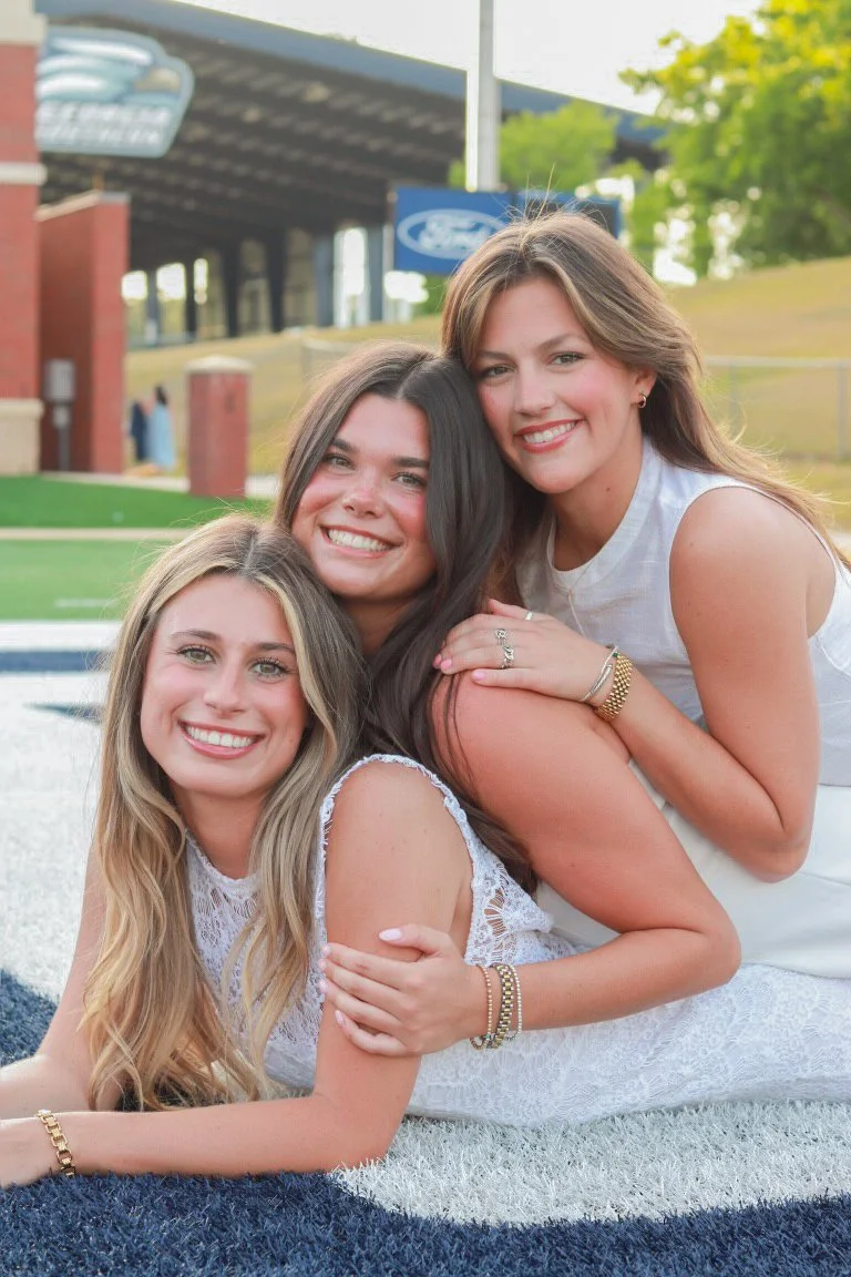 Three young women with light skin sitting and lying on the grass, smiling and posing for a photo outdoors on a sunny day.
