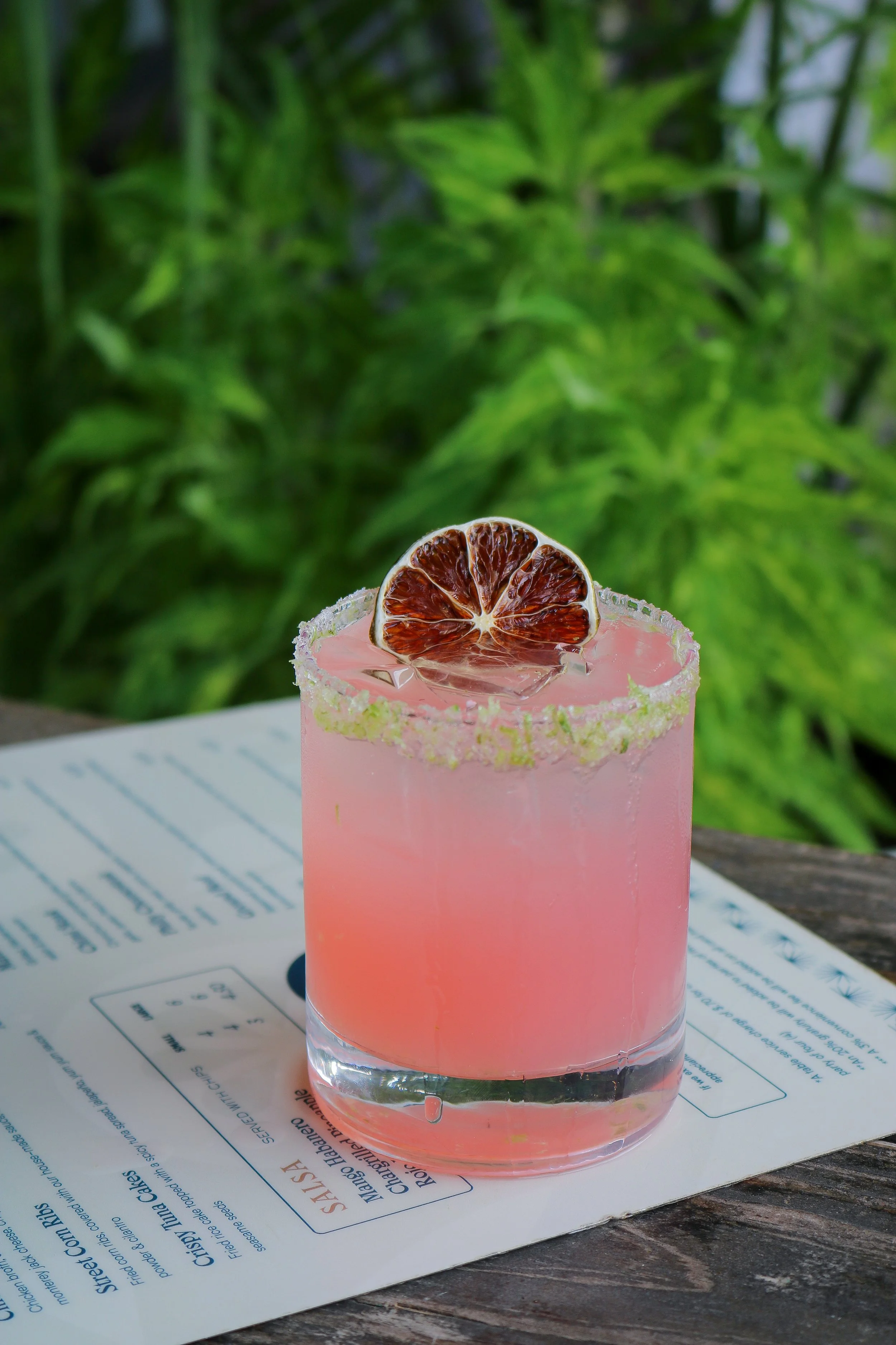 A pink drink in a clear glass with a salted rim, topped with a dried blood orange slice, placed on a paper menu on a wooden surface with green foliage in the background.