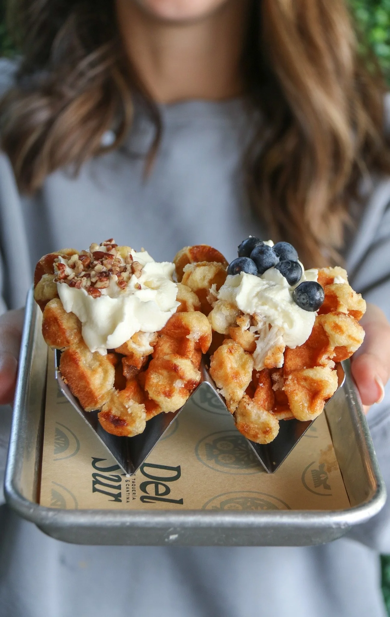 Close-up of a waffle topped with whipped cream, blueberries, and chopped nuts, held in a metal holder over branded paper.