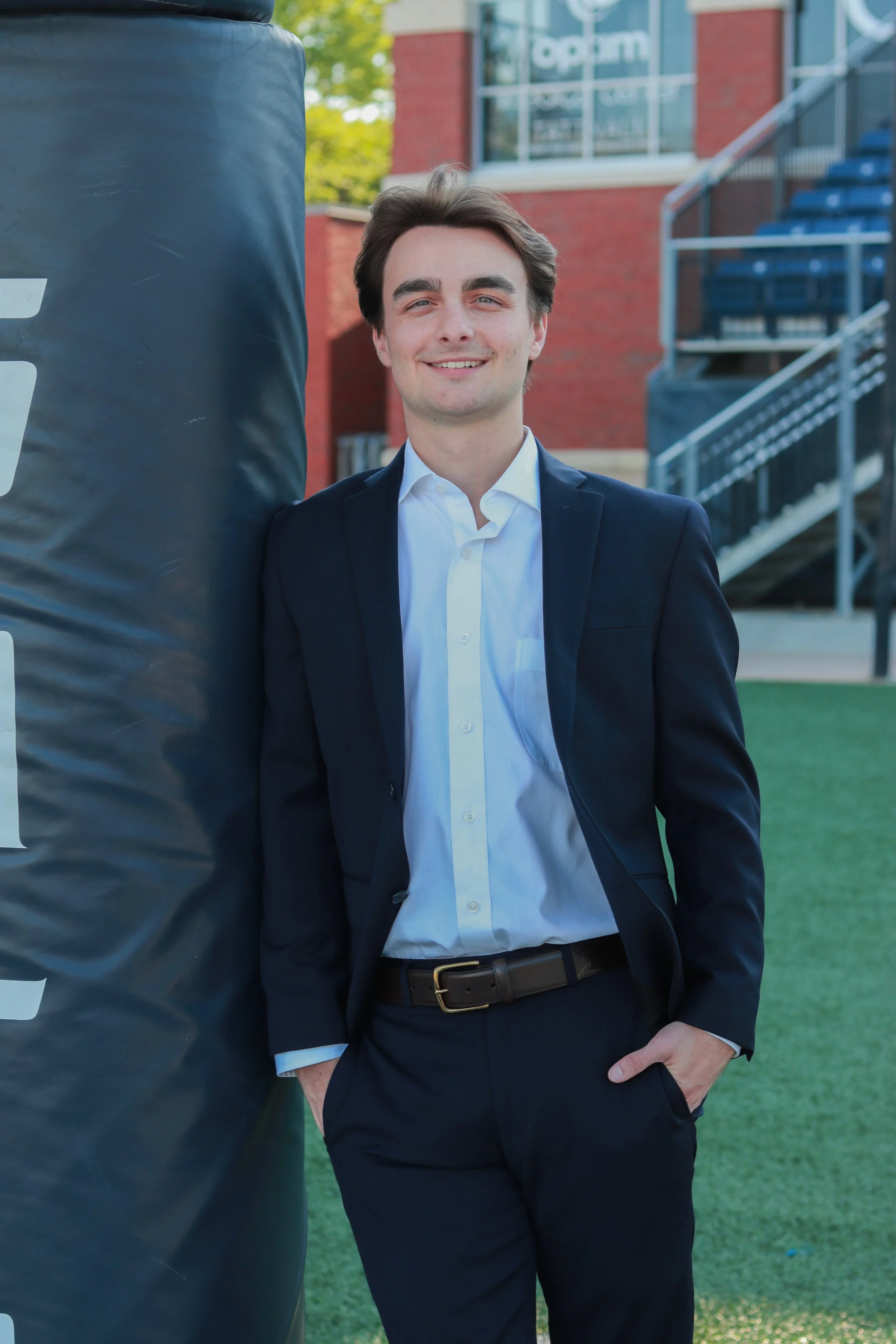 Young man in a suit standing outdoors next to a punching bag, with a brick building and blue bleachers in the background.