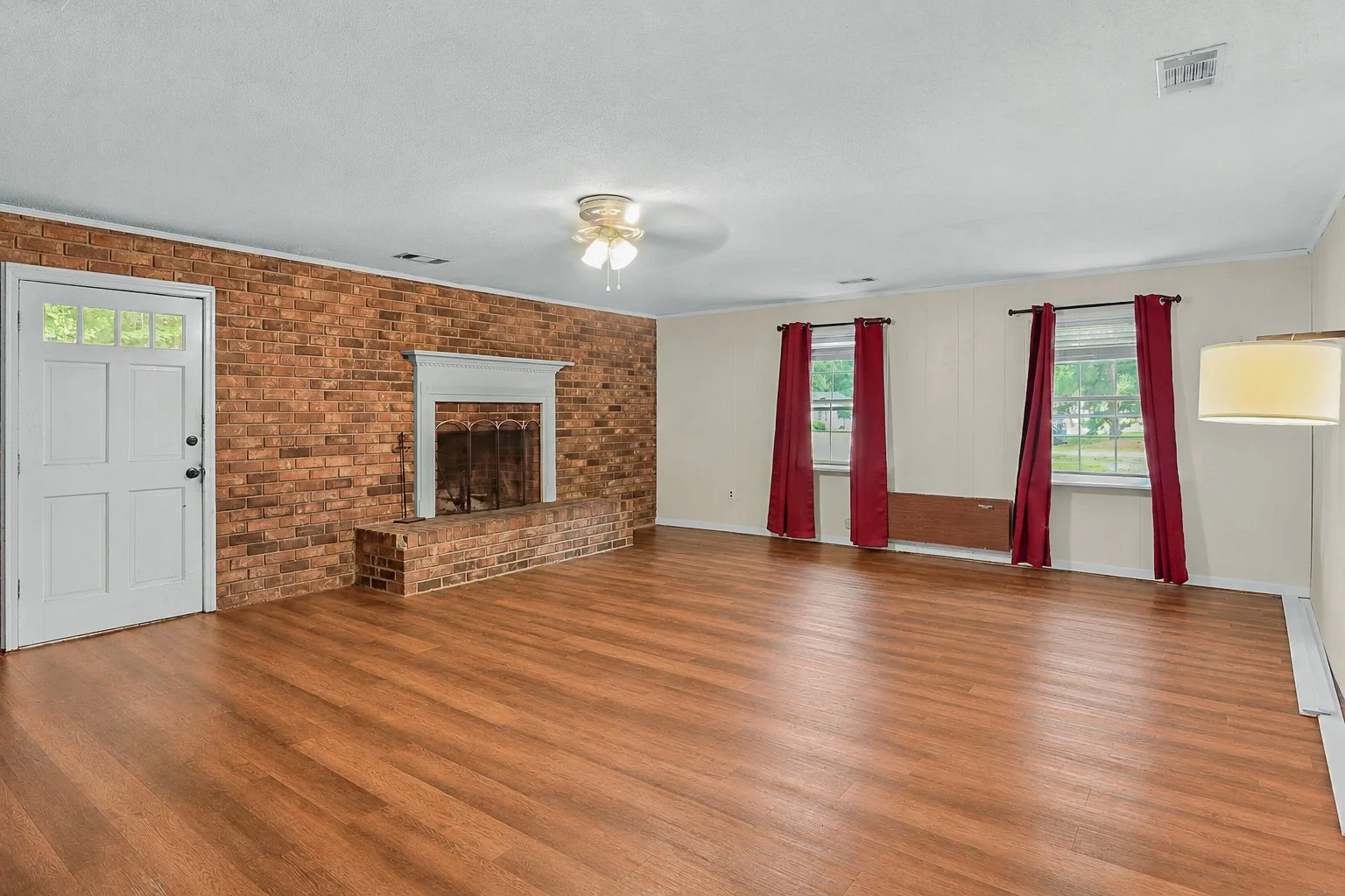 Empty living room with brick fireplace, hardwood floor, and two windows with red curtains.