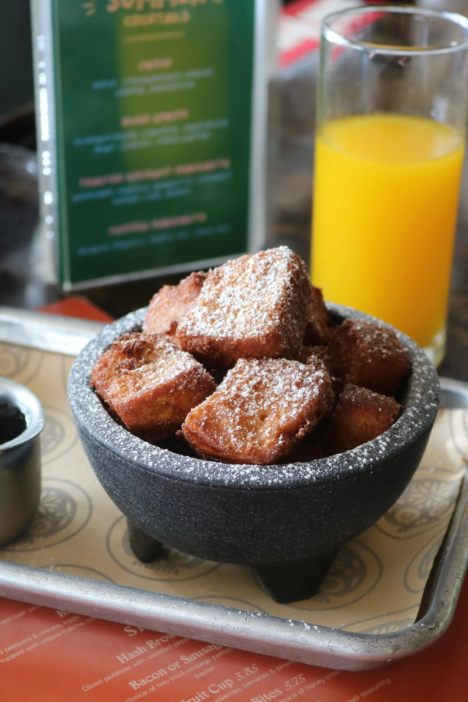 A black stone mortar filled with fried, sugar-dusted cubes of French toast or similar bread, with a glass of orange juice and a blurred menu in the background.