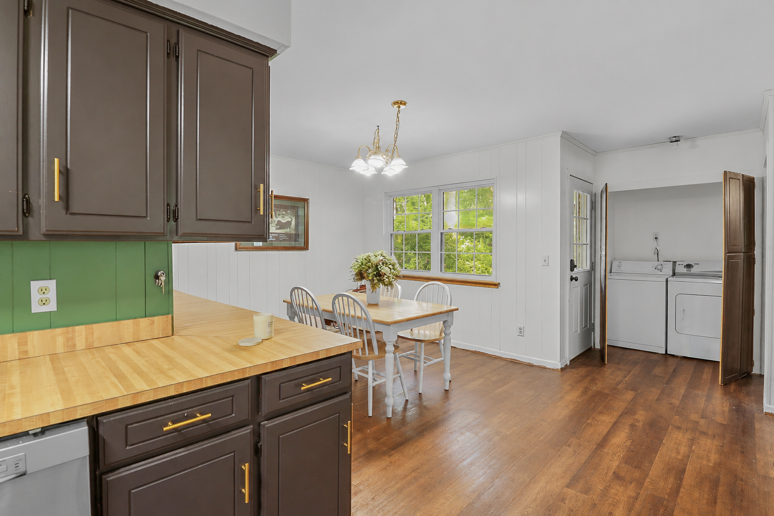 Dining area with a wooden table and four white chairs, a vase of flowers on the table, a window with green trees outside, and a laundry area with a washing machine and dryer in a small room.