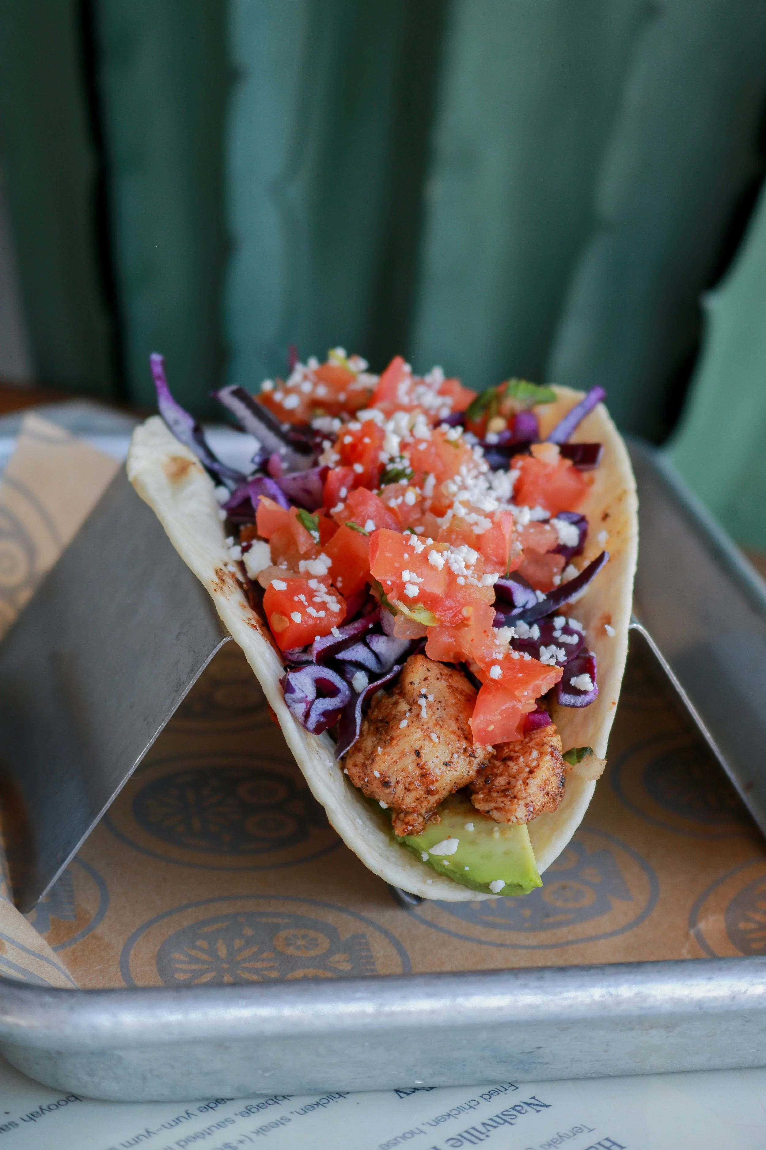 A taco with diced tomatoes, shredded purple cabbage, crumbled cheese, and grilled chicken inside a soft taco shell, held by a hand over a tray.