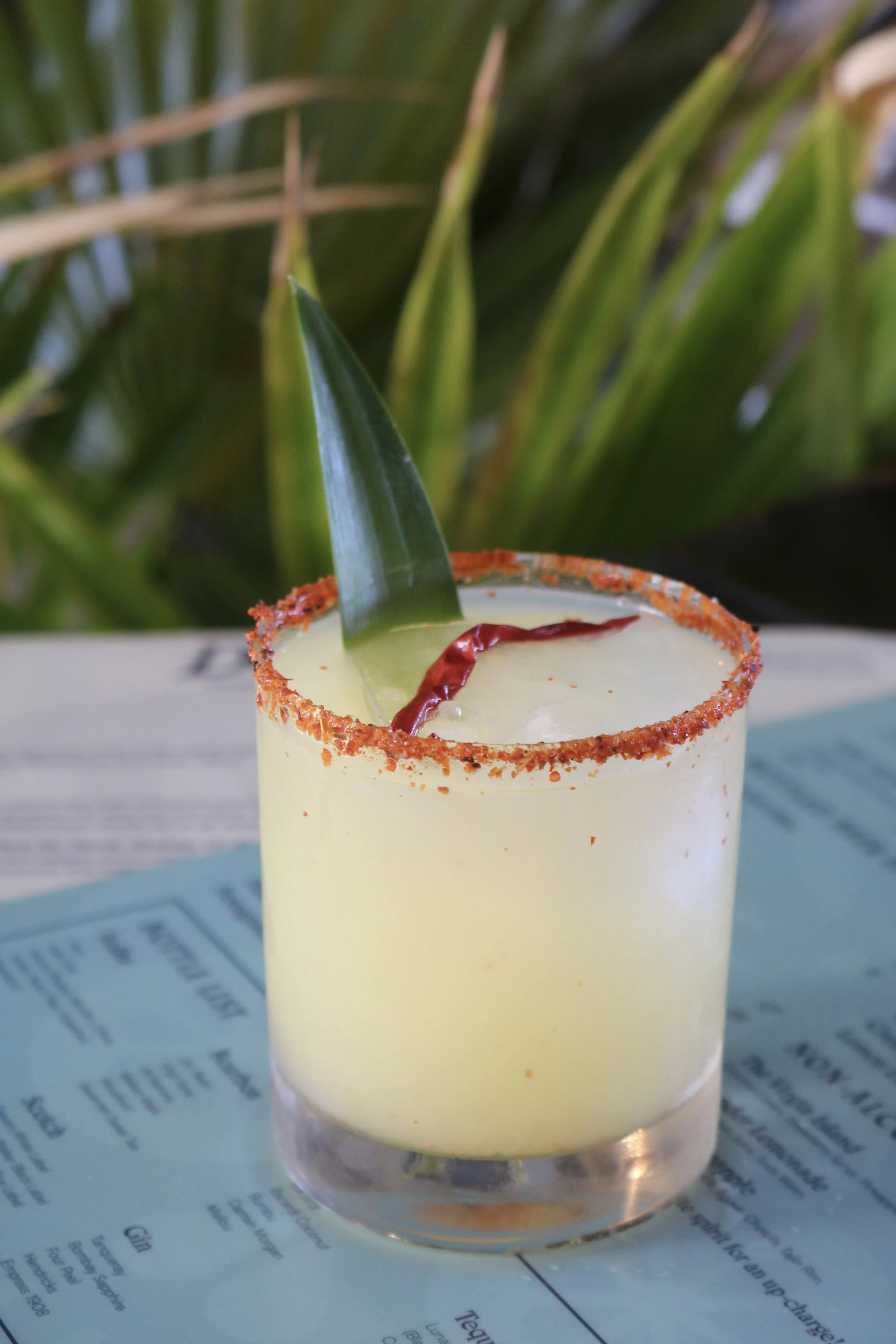 A yellow cocktail drink garnished with a pineapple leaf and a red chili, with a salt or spicy seasoning rim on the glass, placed on a blue sheet of paper.