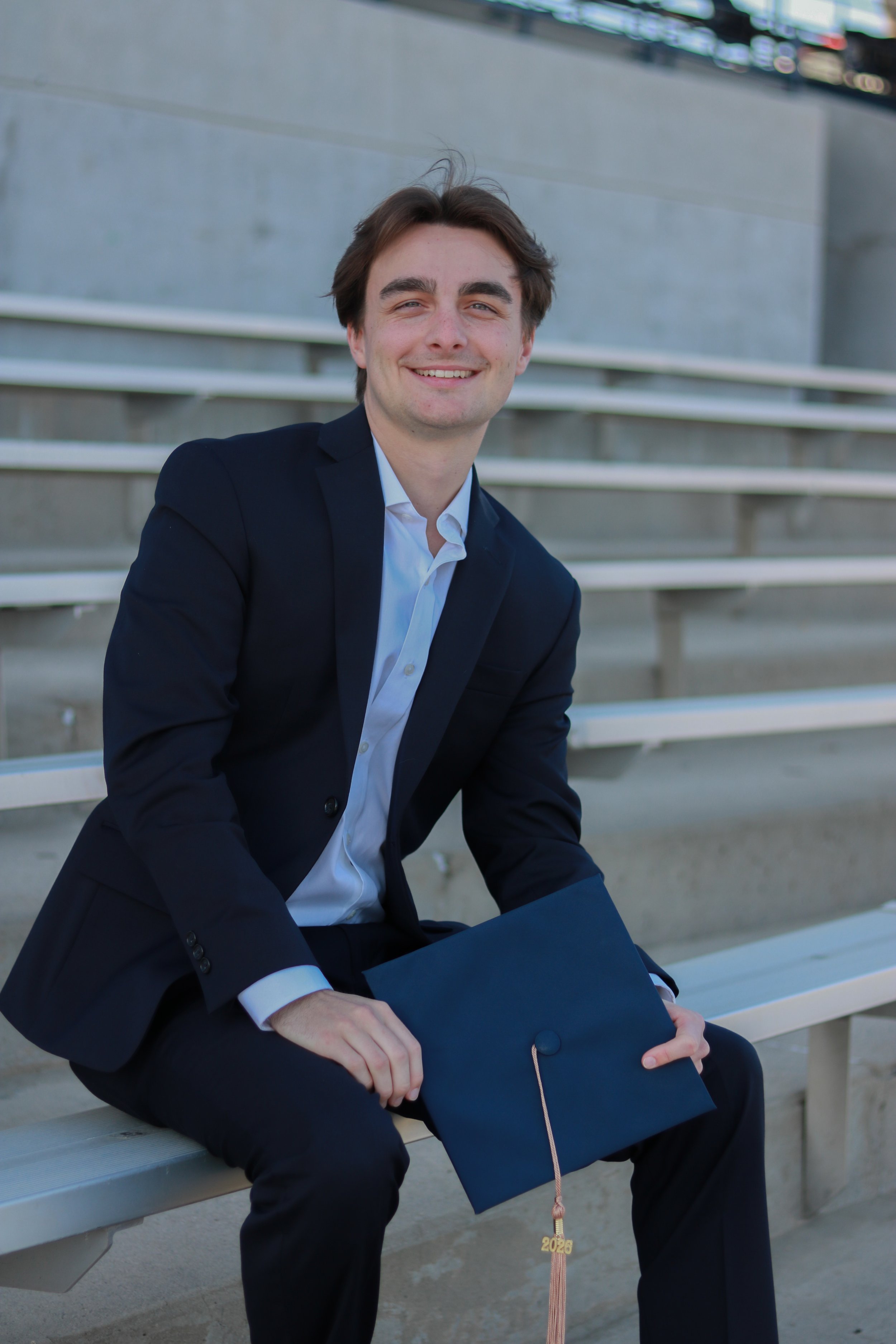 A young man in a dark suit with a light shirt, sitting on a bench, smiling, holding a dark graduation cap and a black gown with a '2020' tassel, with concrete steps and a building in the background.