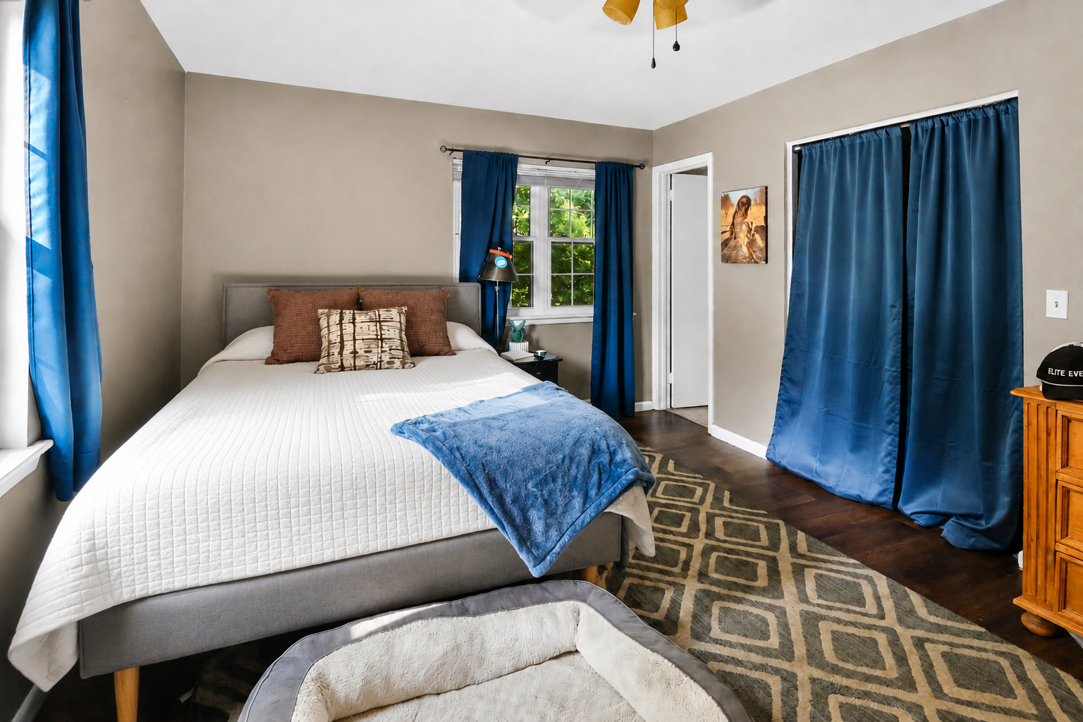 A bedroom with a queen-sized bed, brown pillows, a white quilt, a blue blanket, and a gray headboard. There are curtains on the windows, one black lamp, a small planner, and a framed picture on the wall. The floor is hardwood with a patterned area ru