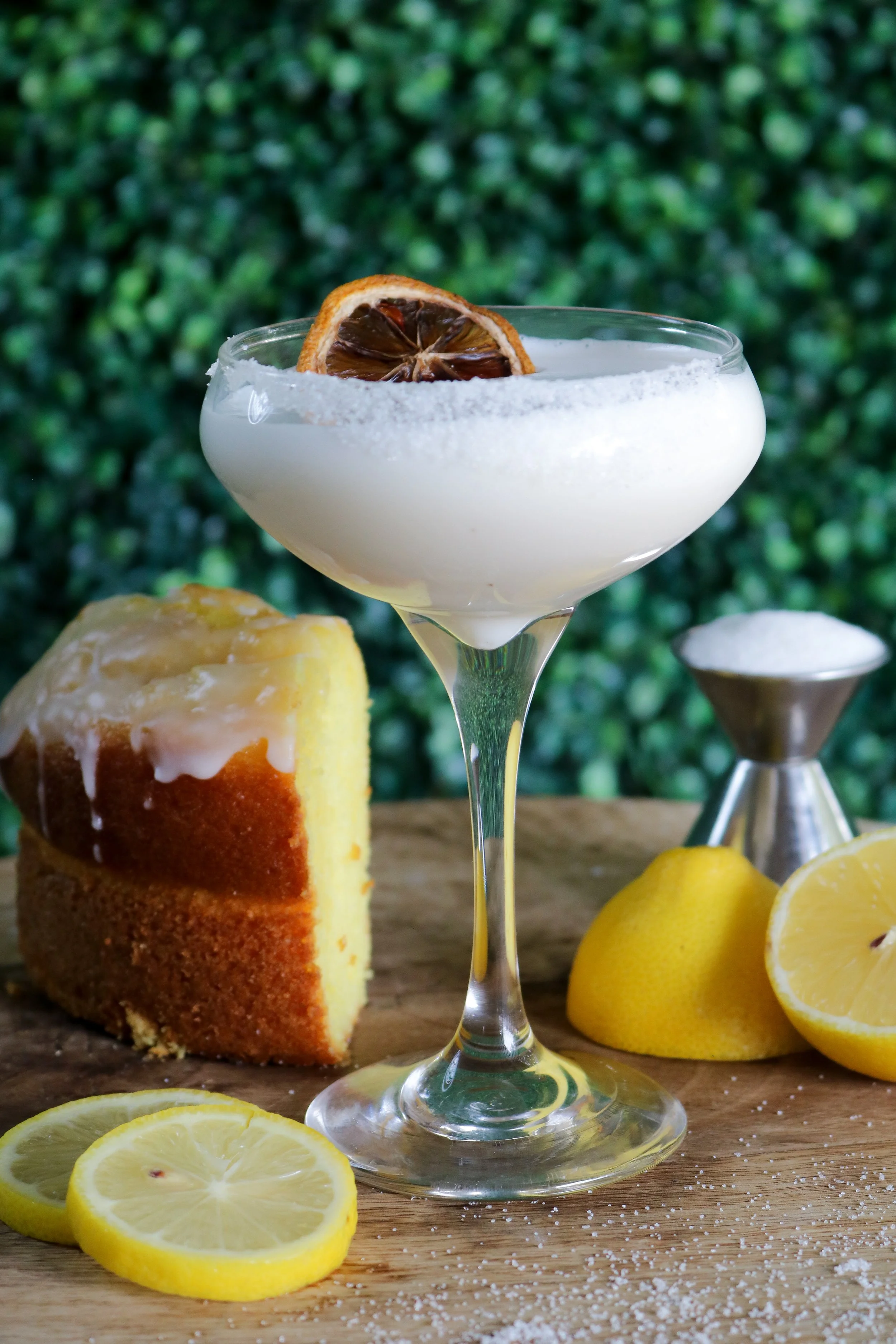A glass of lemon cocktail garnished with dried lemon slices, surrounded by fresh lemon slices, a slice of lemon cake with icing, and scattered coconut flakes on a wooden surface with a green leafy background.