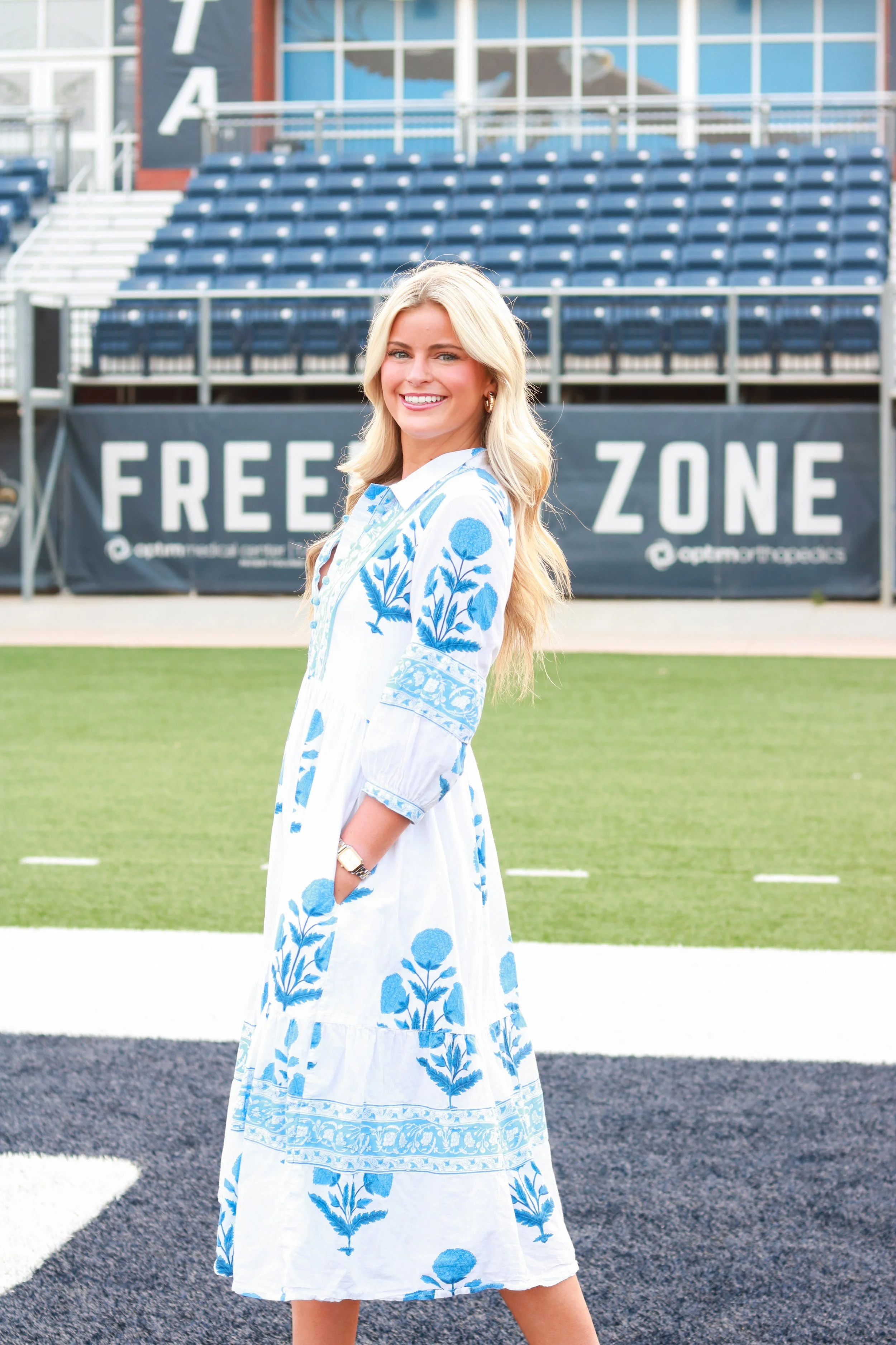 A blonde woman in a white dress with blue floral embroidery standing on a sports field with empty stadium seats in the background.