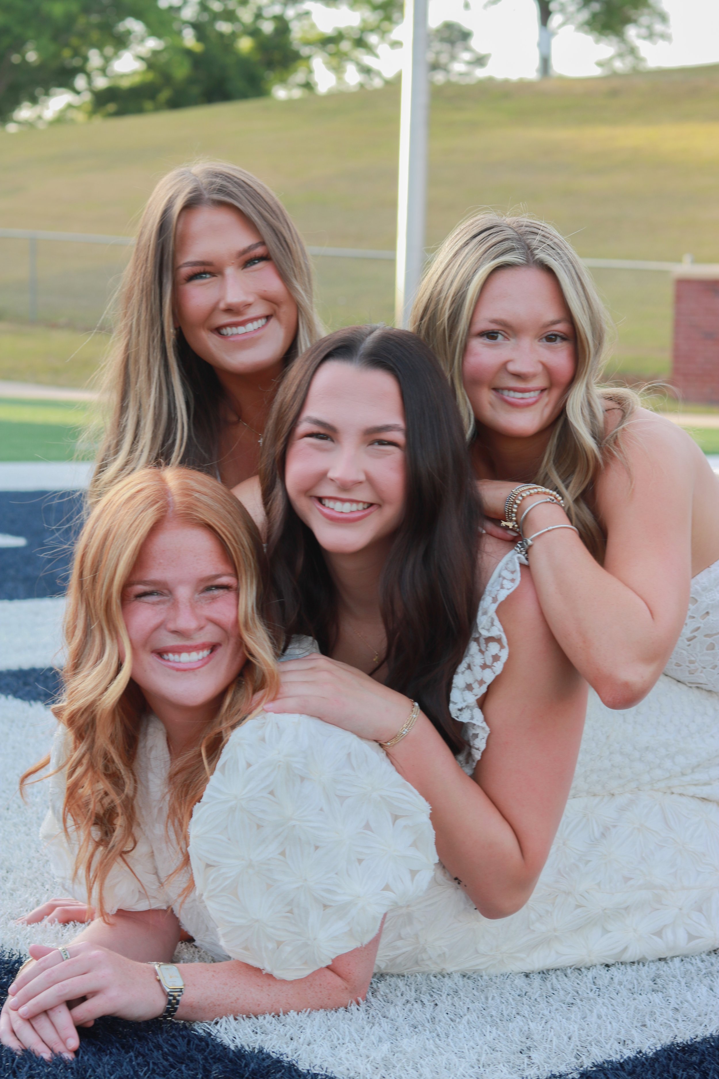 Group of six women lying on the grass outdoors, smiling and posing together. They are dressed in light-colored dresses and are surrounded by trees and a grassy hill.