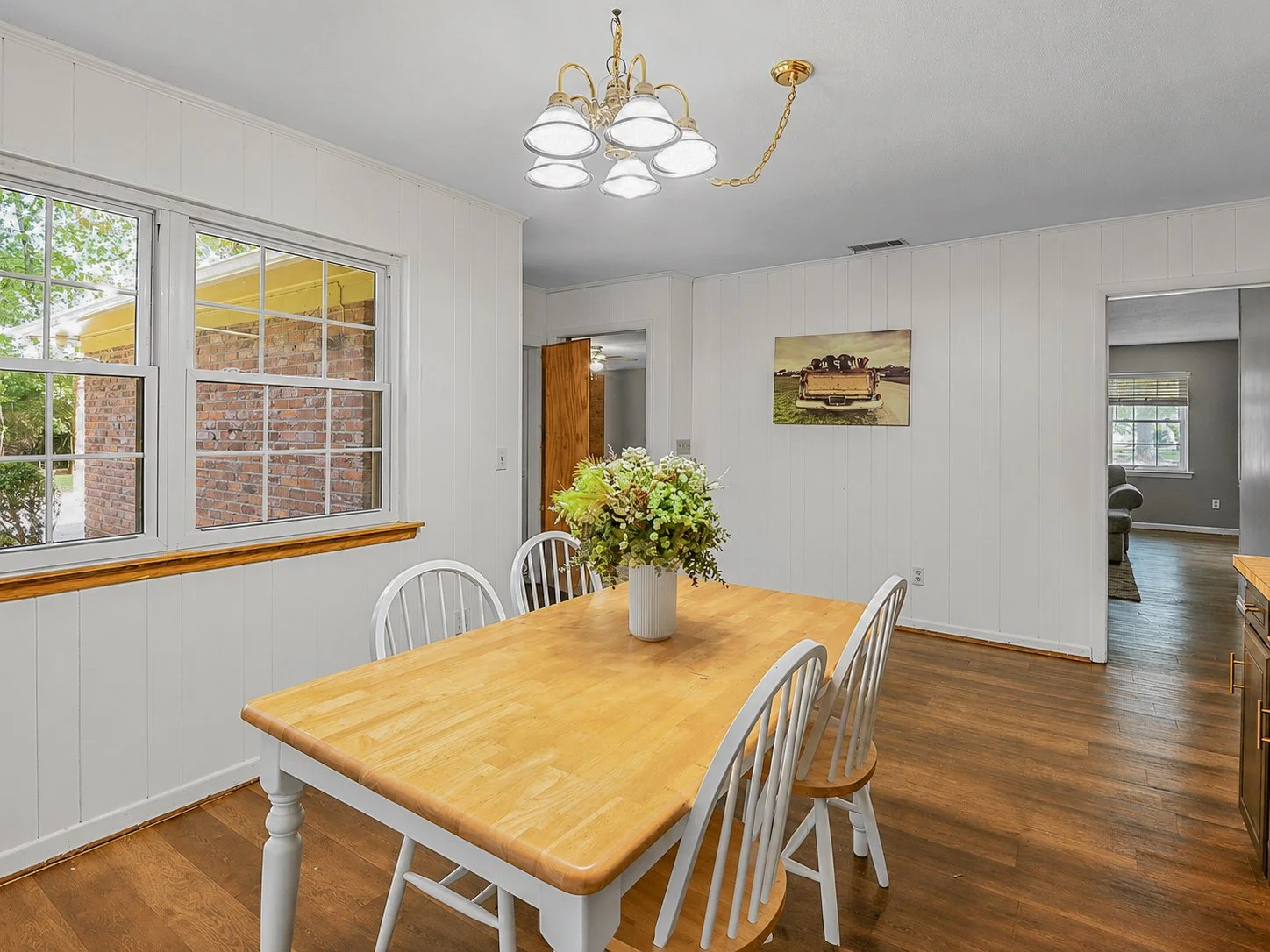 A dining room with a wooden table and six white chairs, a vase with flowers on the table, large windows showing an outside brick wall and trees, a ceiling light fixture, and a doorway leading to a living room with a window and couch.