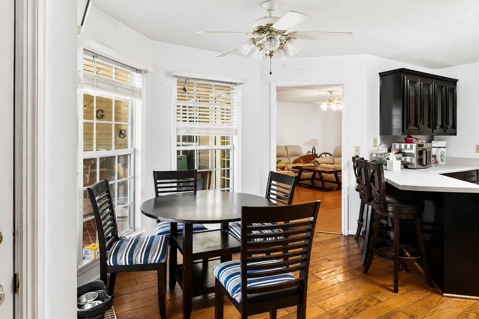 Dining area with round table, black chairs with striped cushions, next to glass door leading to outdoor porch, view into living room with beige sofas and wooden coffee table, kitchen with black cabinets and bar stools.