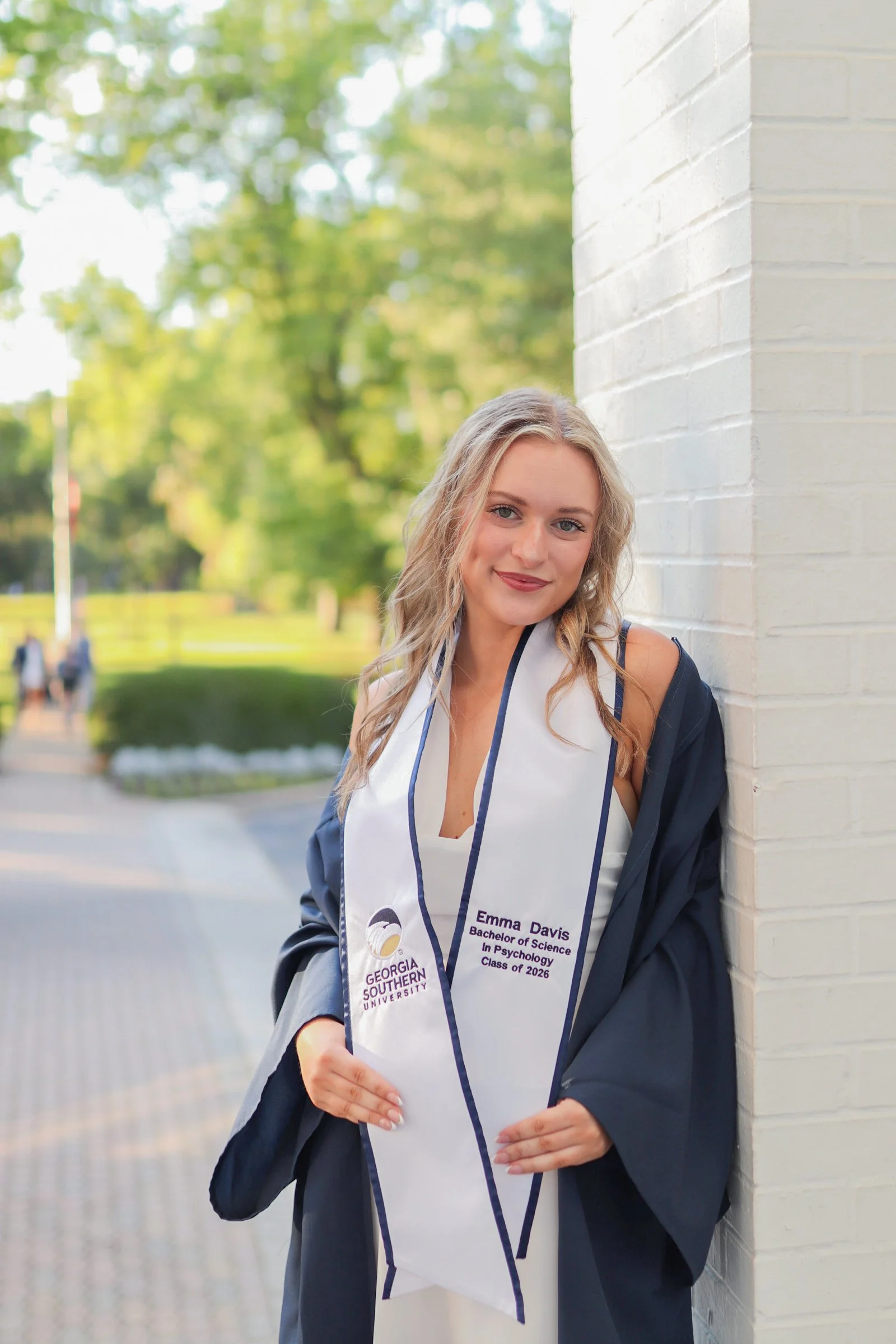 Young woman in graduation cap and gown leaning against a white brick wall outdoors, holding her diploma and smiling.