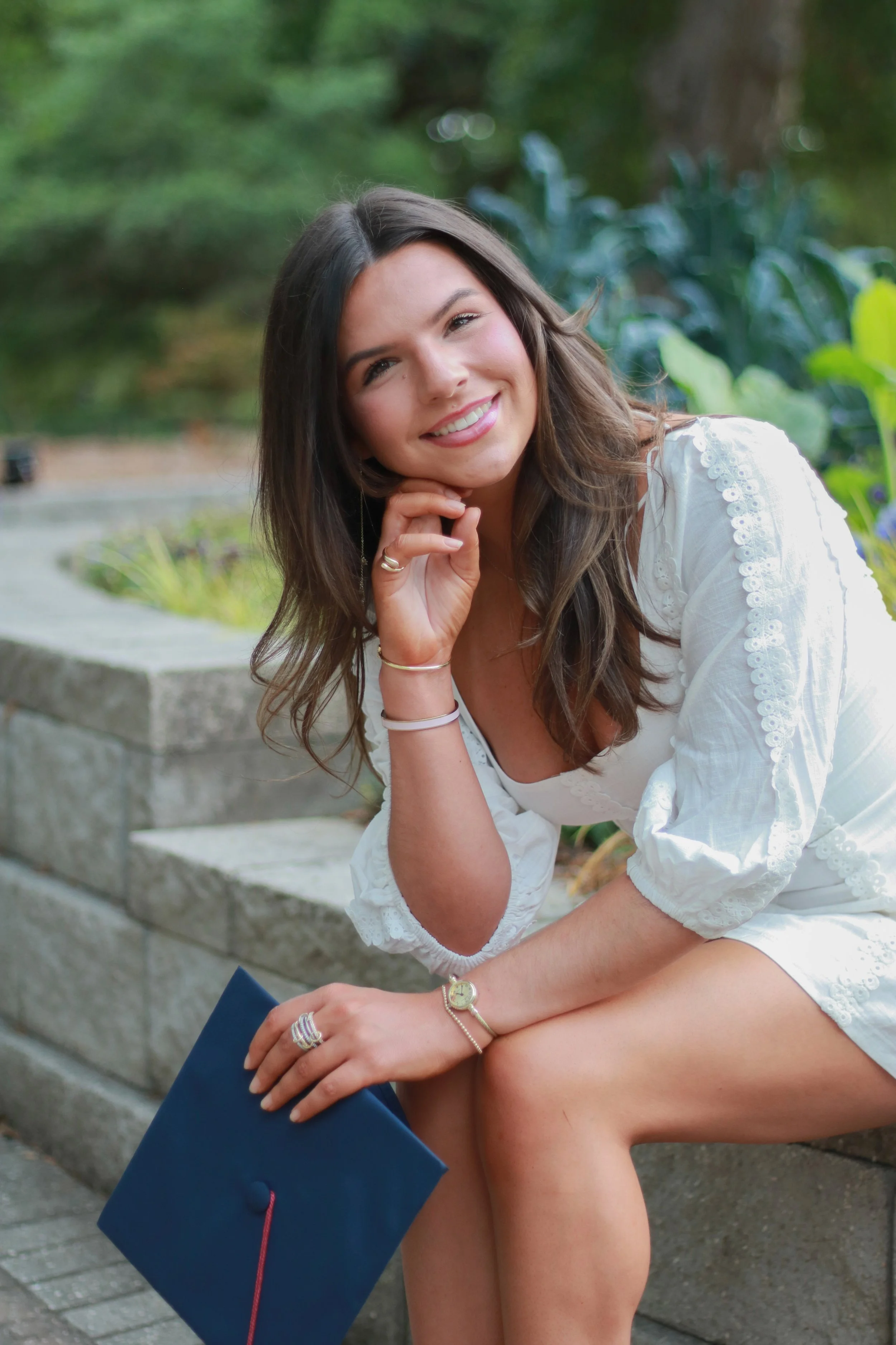 A young woman with long brown hair, wearing a white lace dress, smiling while resting her chin on her hand, sitting outdoors on stone steps surrounded by greenery. She holds a blue folder with a pink ribbon, wearing jewelry including rings, a bracele