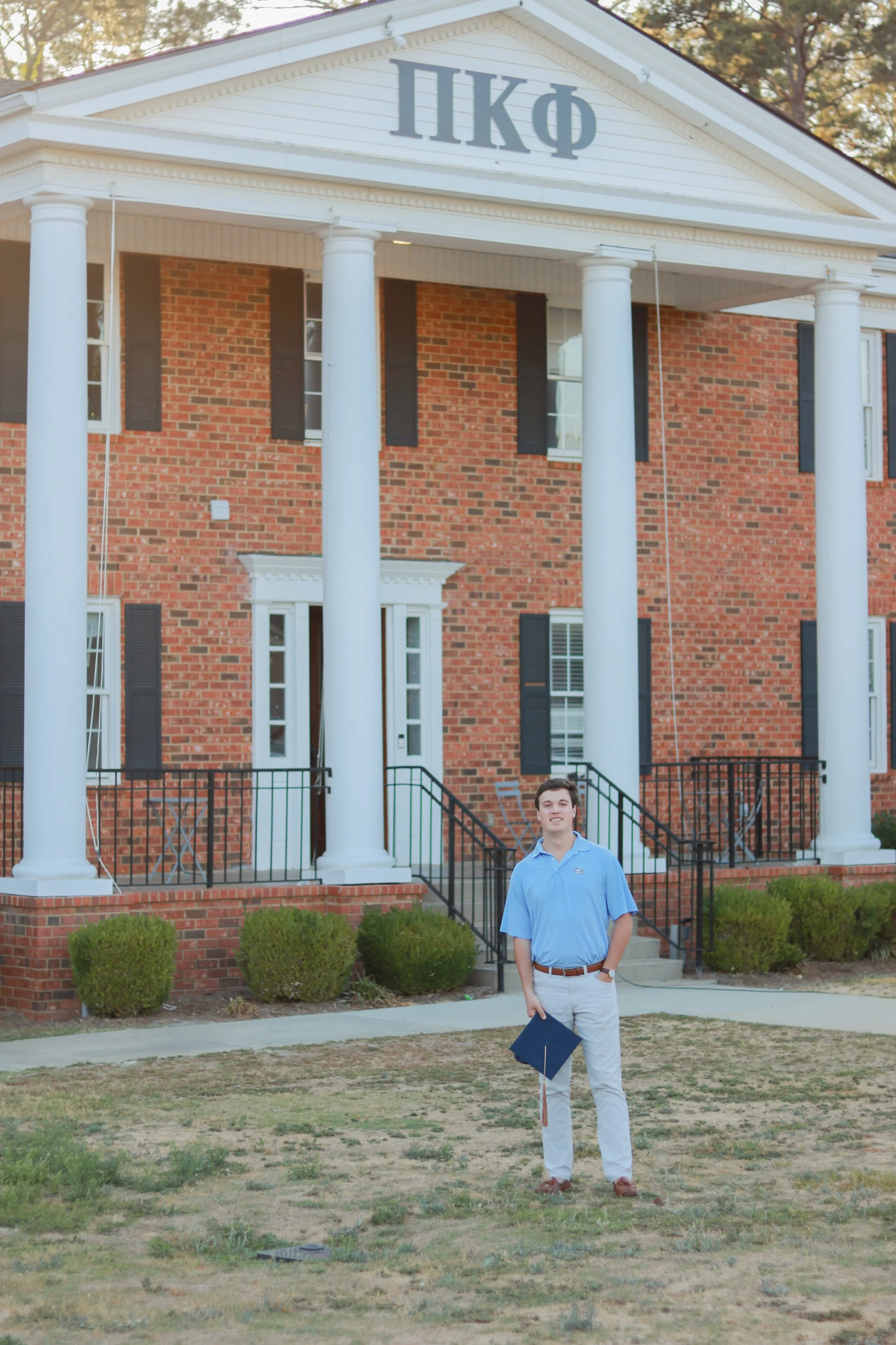 A young man in a light blue shirt and tan pants standing on a grassy lawn, holding a graduation cap and smiling in front of a large brick building with white columns, black shutters, and Greek letters on the pediment.