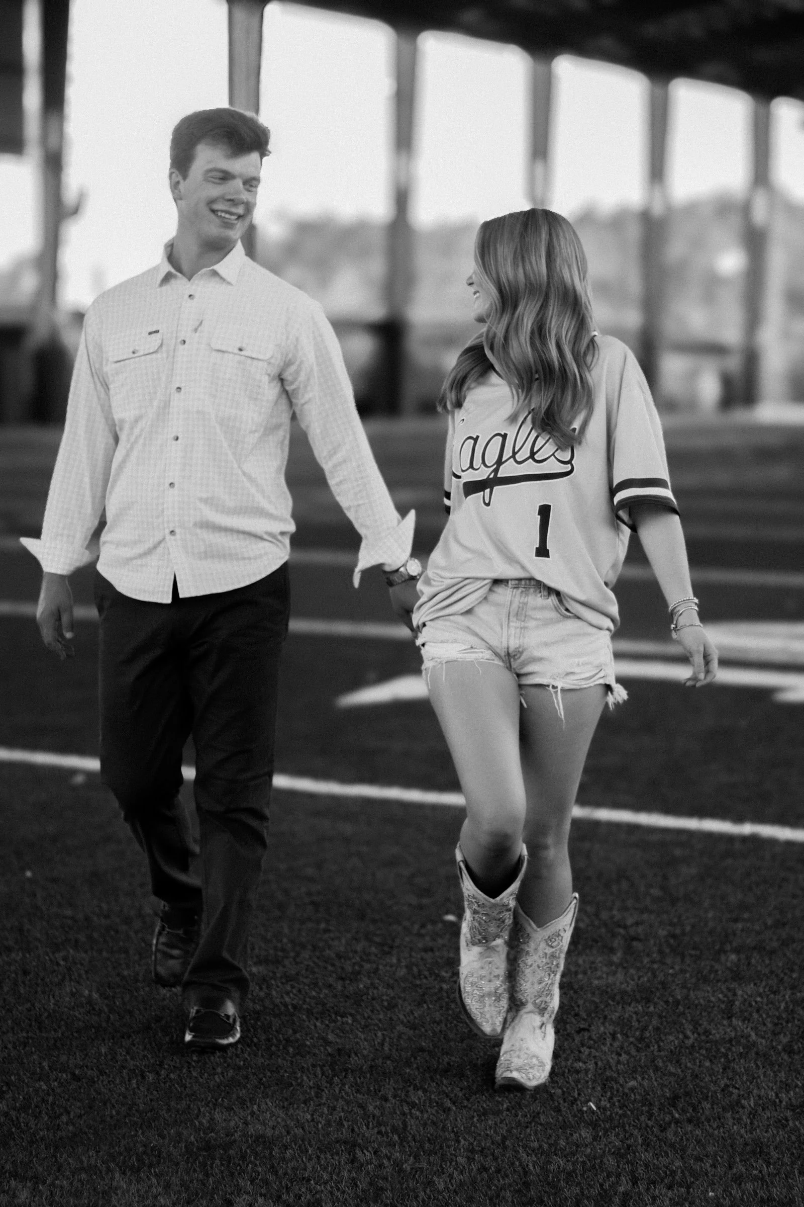 A young man and young woman walking together on a football field, smiling and holding hands. The woman is wearing a sports jersey and cowboy boots, and the man is dressed in a button-up shirt and dark pants.