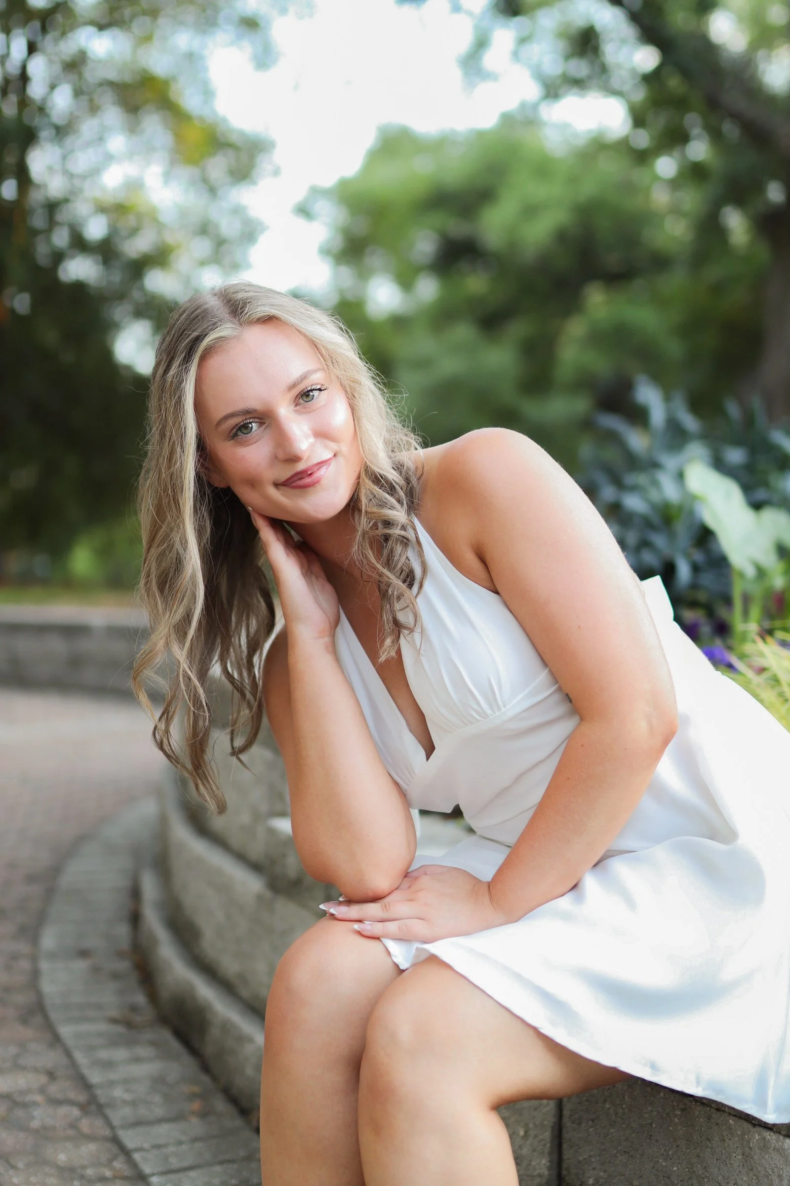 A smiling woman with wavy blonde hair wearing a white sleeveless dress, sitting on a stone bench in a park with green trees in the background, resting her head on her hand.