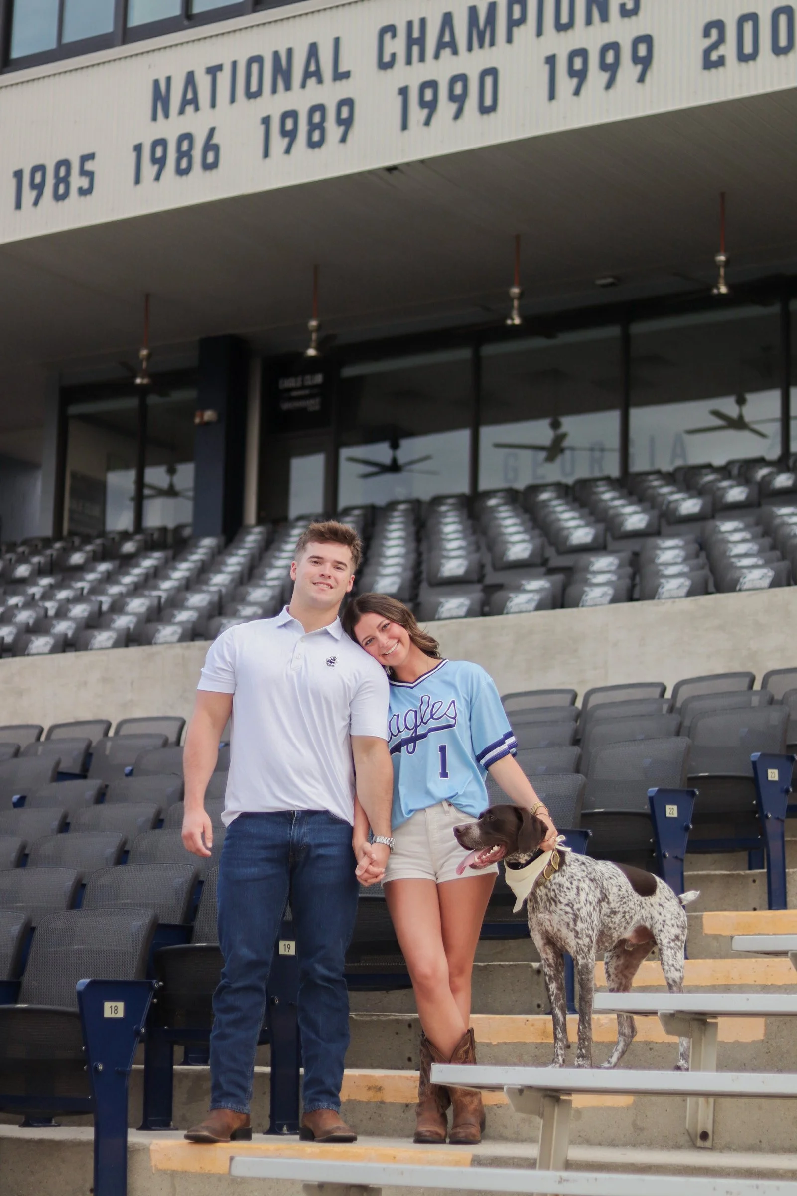 A smiling young couple with their German Shorthaired Pointer dog while standing on the steps of a baseball stadium with empty spectator seats in the background. The woman is wearing a blue sports jersey, and the man is in a white polo shirt with jean