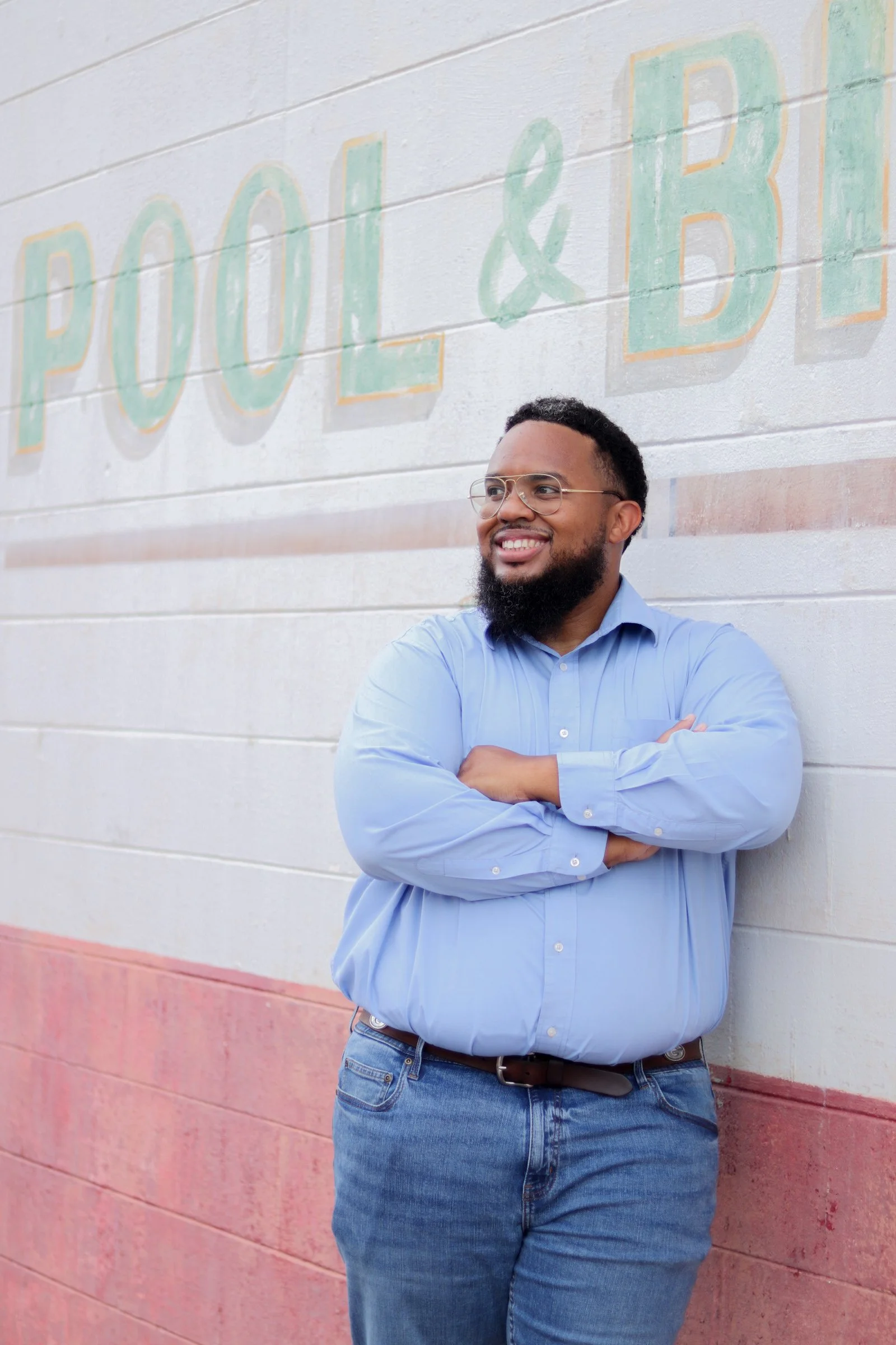 A man with glasses and a beard, wearing a light blue dress shirt and jeans, standing with arms crossed in front of a colorful painted brick wall with the words 'POOL & B' visible.