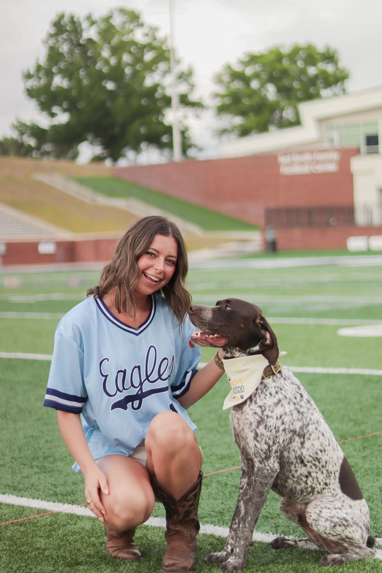 A young woman in a light blue cheerleading uniform smiling and kneeling next to a large, spotted dog on a football field. The dog wears a bandana. In the background, there are trees and a building with a sign.