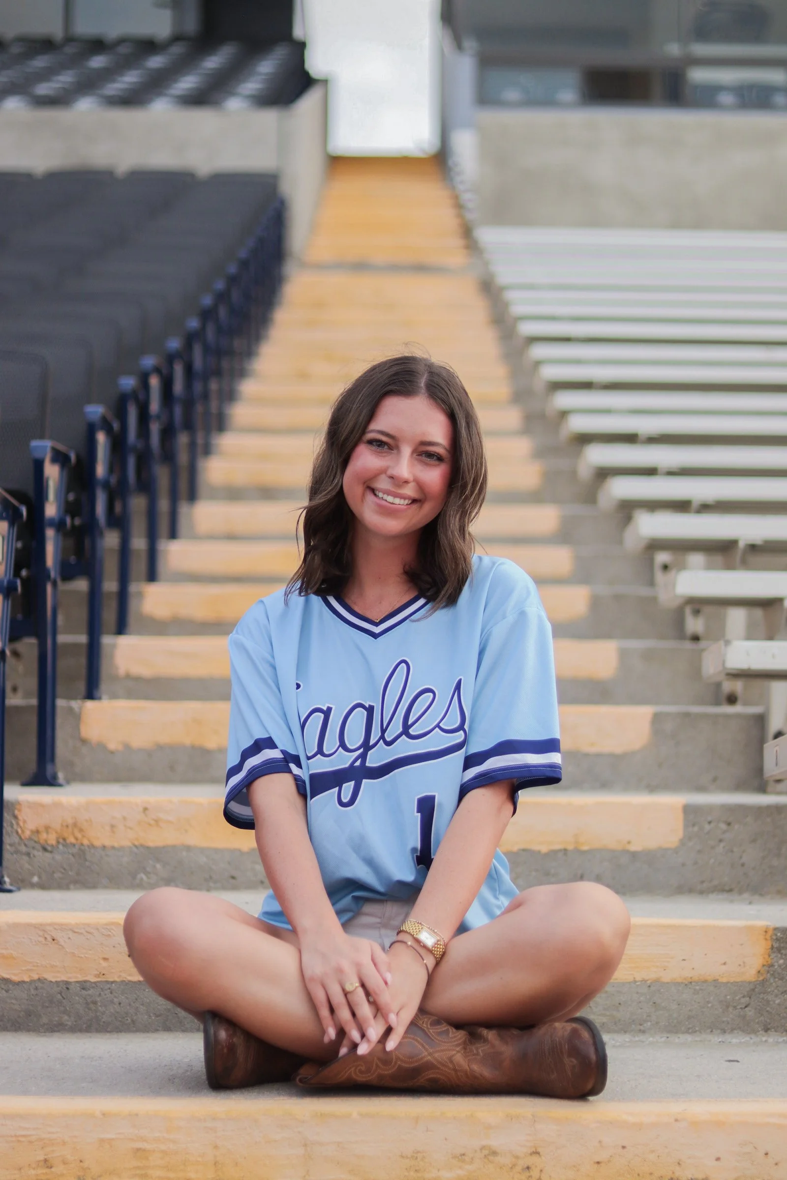 A young woman sitting on outdoor stadium steps, smiling, wearing a light blue sports jersey with 'Eagles' written on it, beige shorts, and brown cowboy boots.