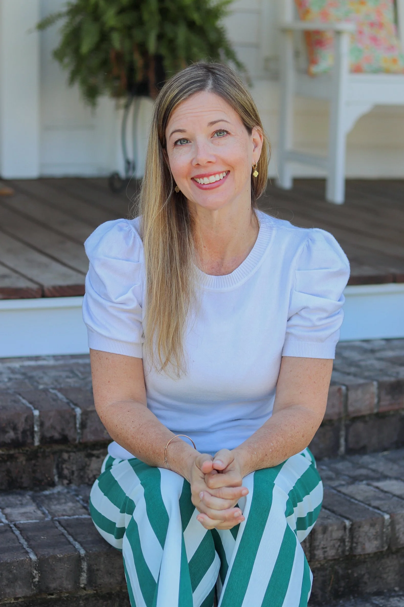 A woman with long blonde hair, wearing a white top and green-and-white striped pants, sitting on brick stairs in front of a background with a potted plant and a white furniture piece with floral cushions, smiling at the camera.