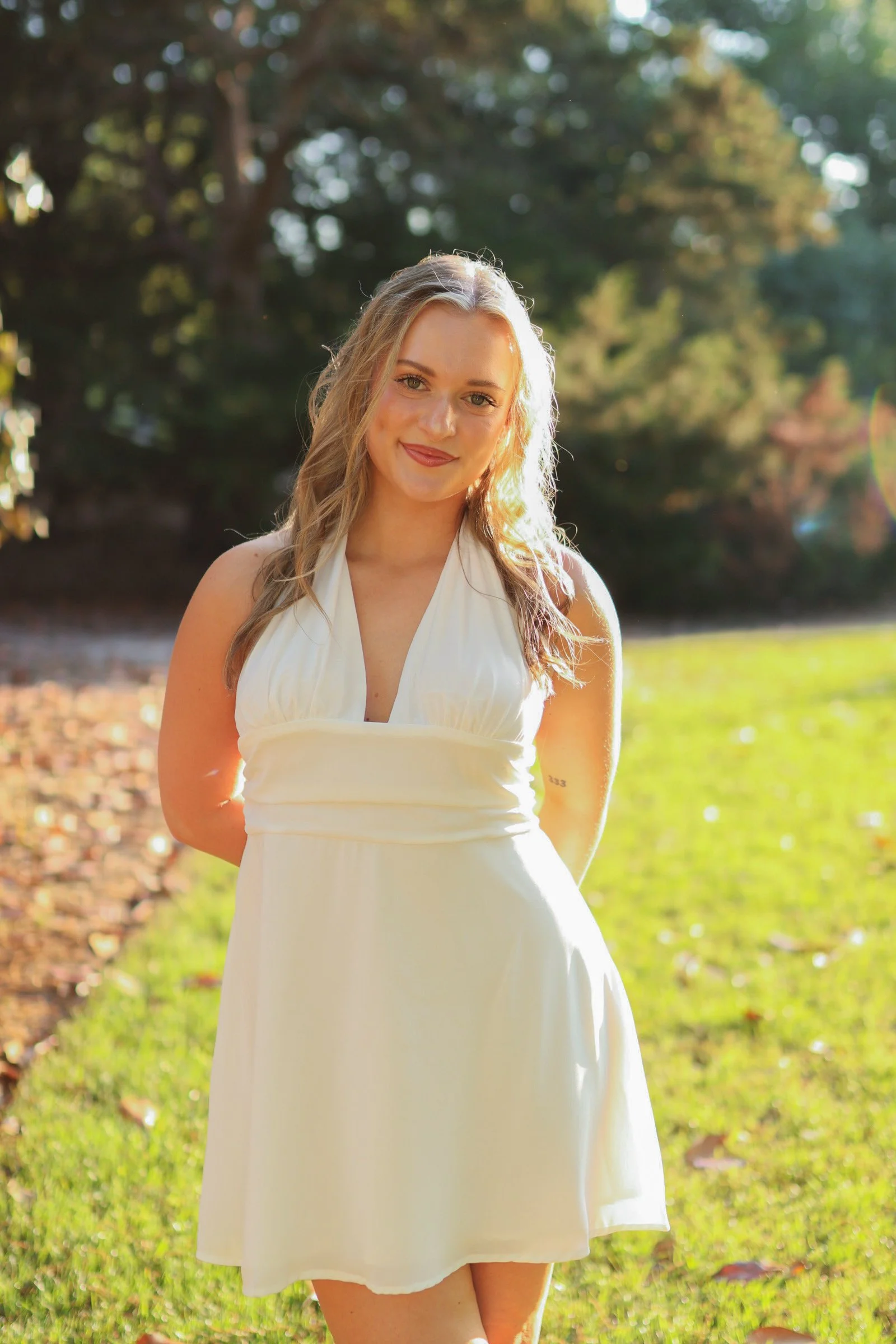 A young woman with blonde hair and fair skin standing outdoors in a grassy area with trees in the background, smiling at the camera, wearing a white sleeveless dress.
