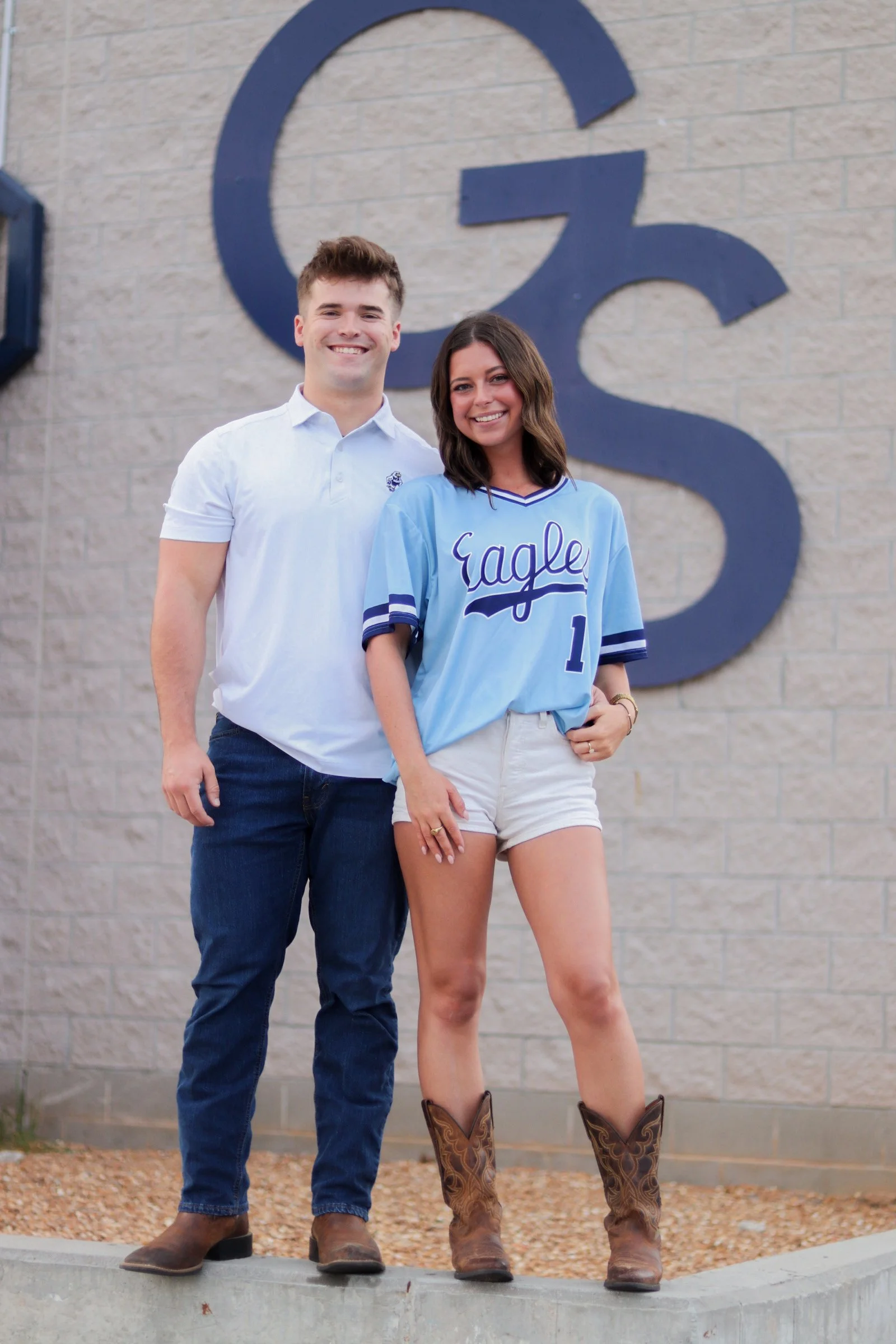 A young man and woman standing together outside, smiling at the camera, with a large letter 'G' in the background, both in casual clothing and cowboy boots.